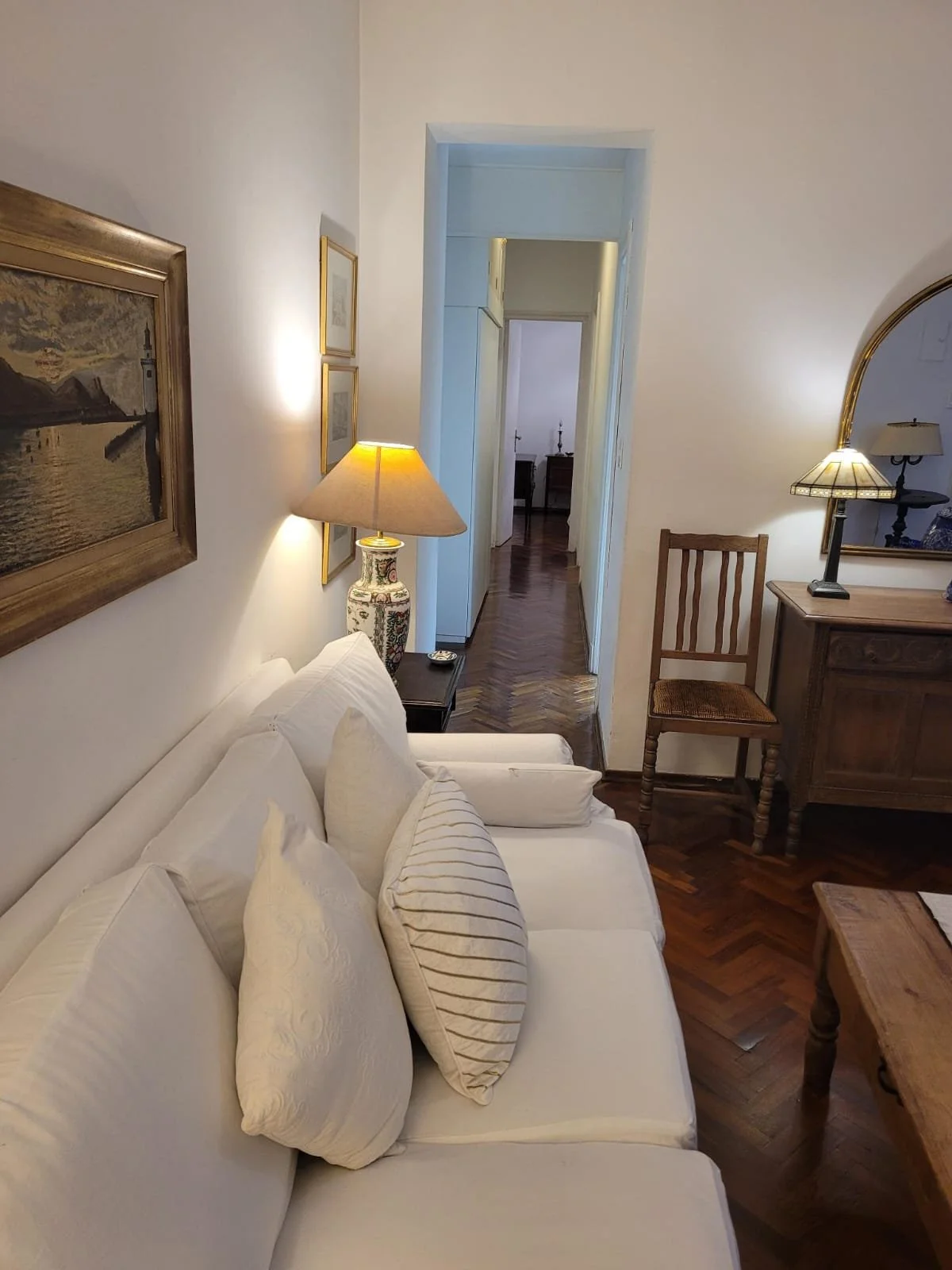 Living room with white couch, wooden table, chairs, lamps, wall art, and hardwood floors.