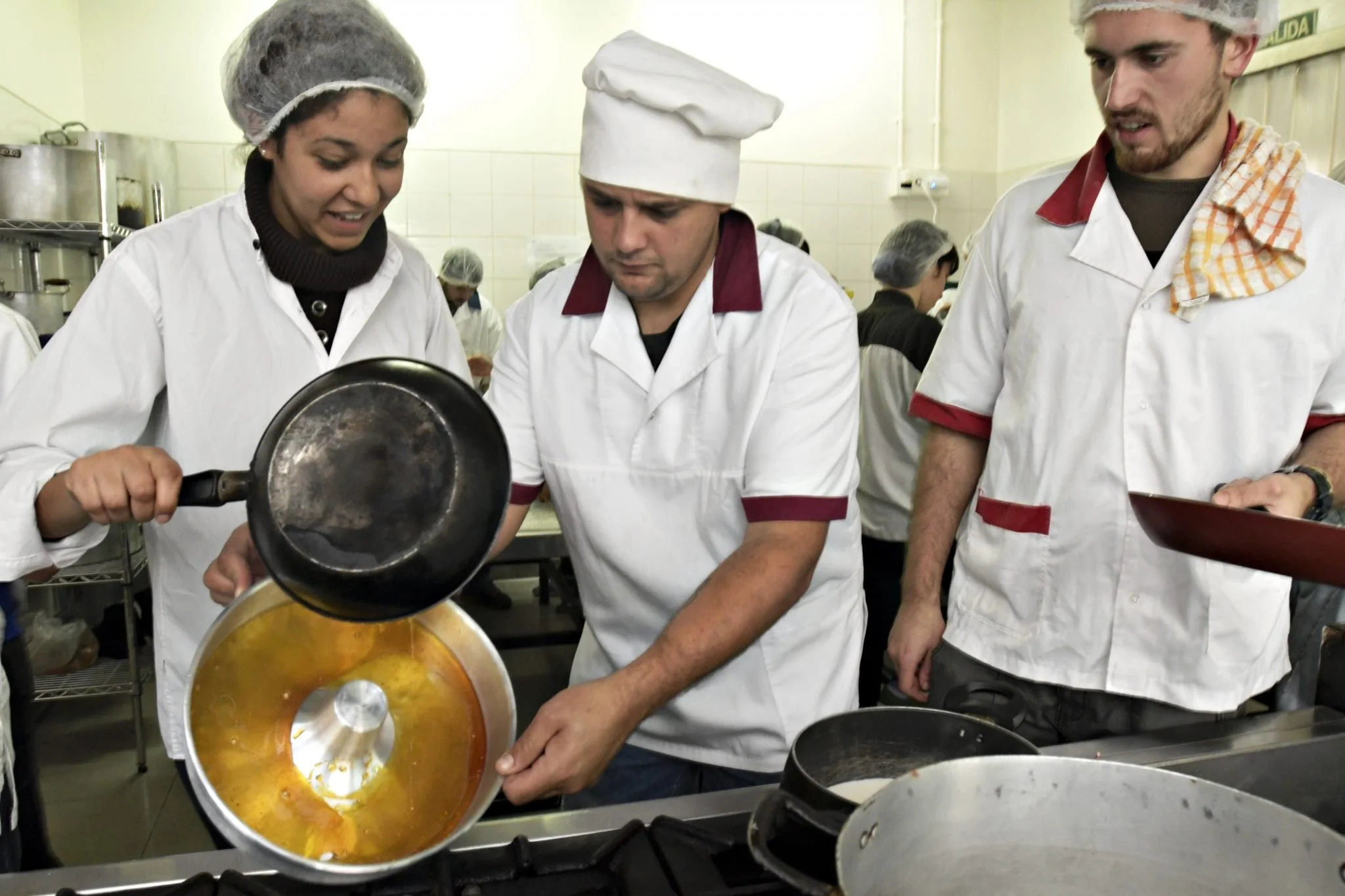 People in a commercial kitchen wearing white uniforms and hairnets, cooking and preparing food.