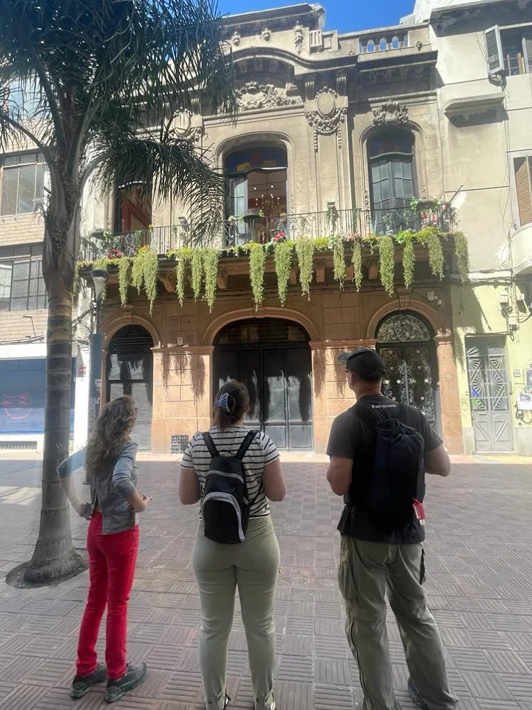 Three people standing on a city sidewalk in front of a historic building with ornate architectural details and flower boxes, with a palm tree on the left.