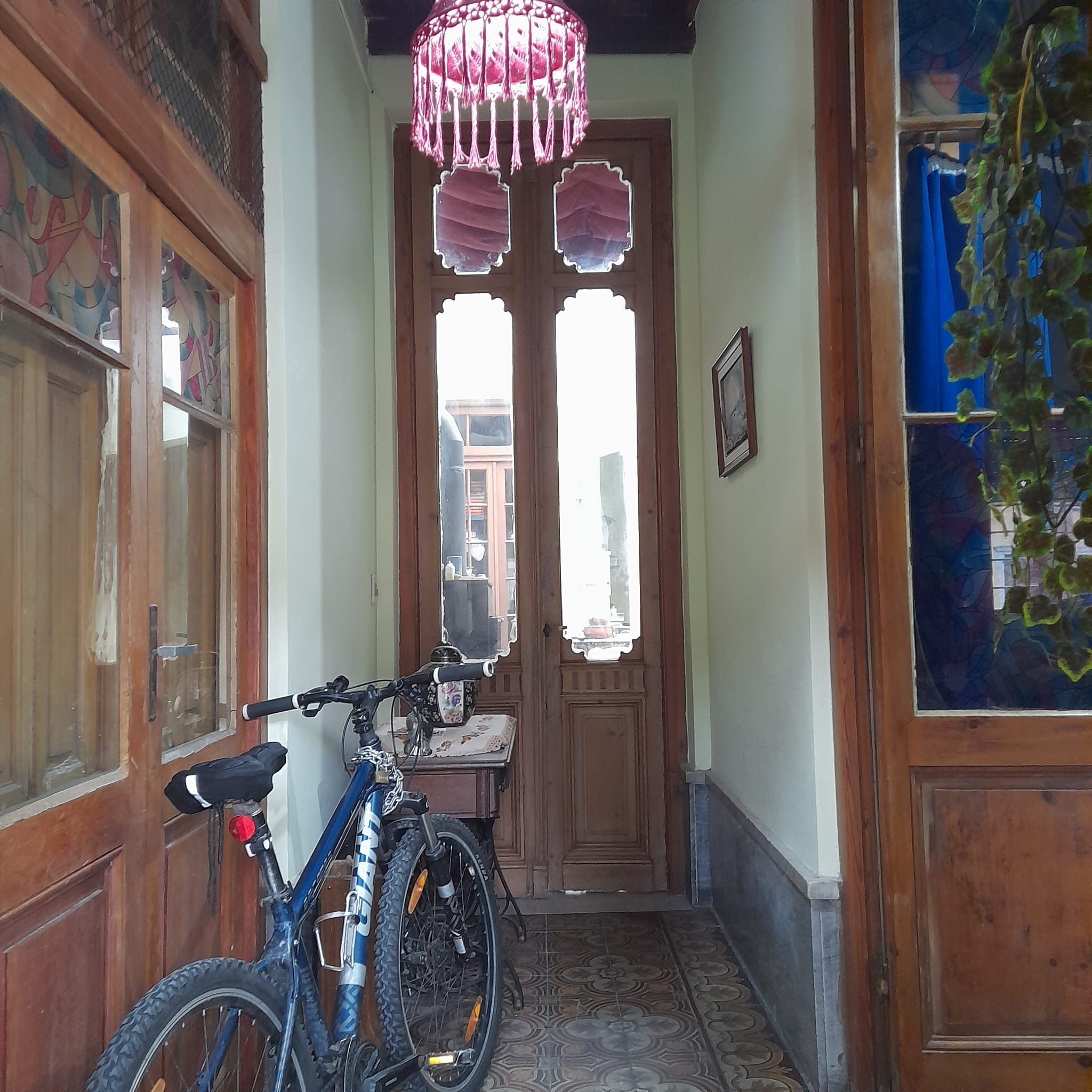 Indoor hallway with wooden doors and windows, a blue bicycle leaning against the left wall, stained glass window, pink chandelier hanging from ceiling, framed picture on the right wall, patterned tile floor, and a small table with a decorative cloth.