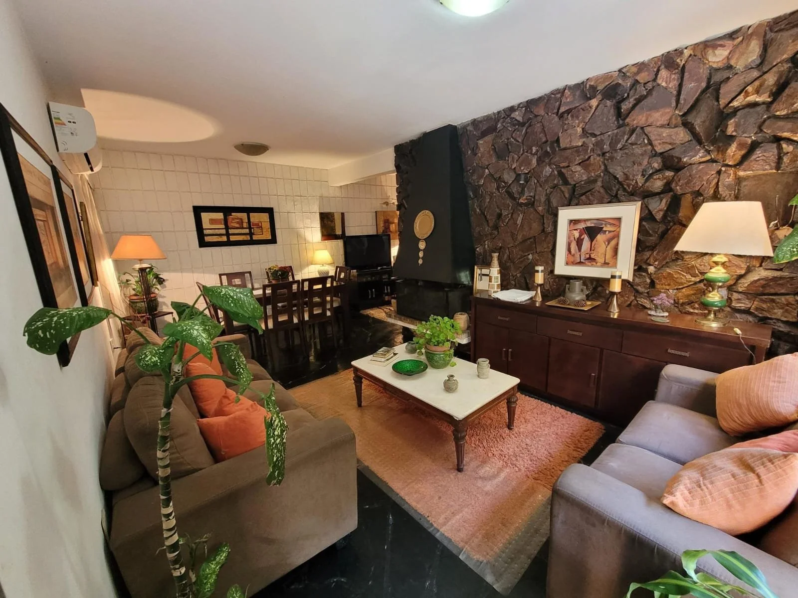 Living room with sofas, coffee table, fireplace, dining area, plants, lamps, and framed art on walls.