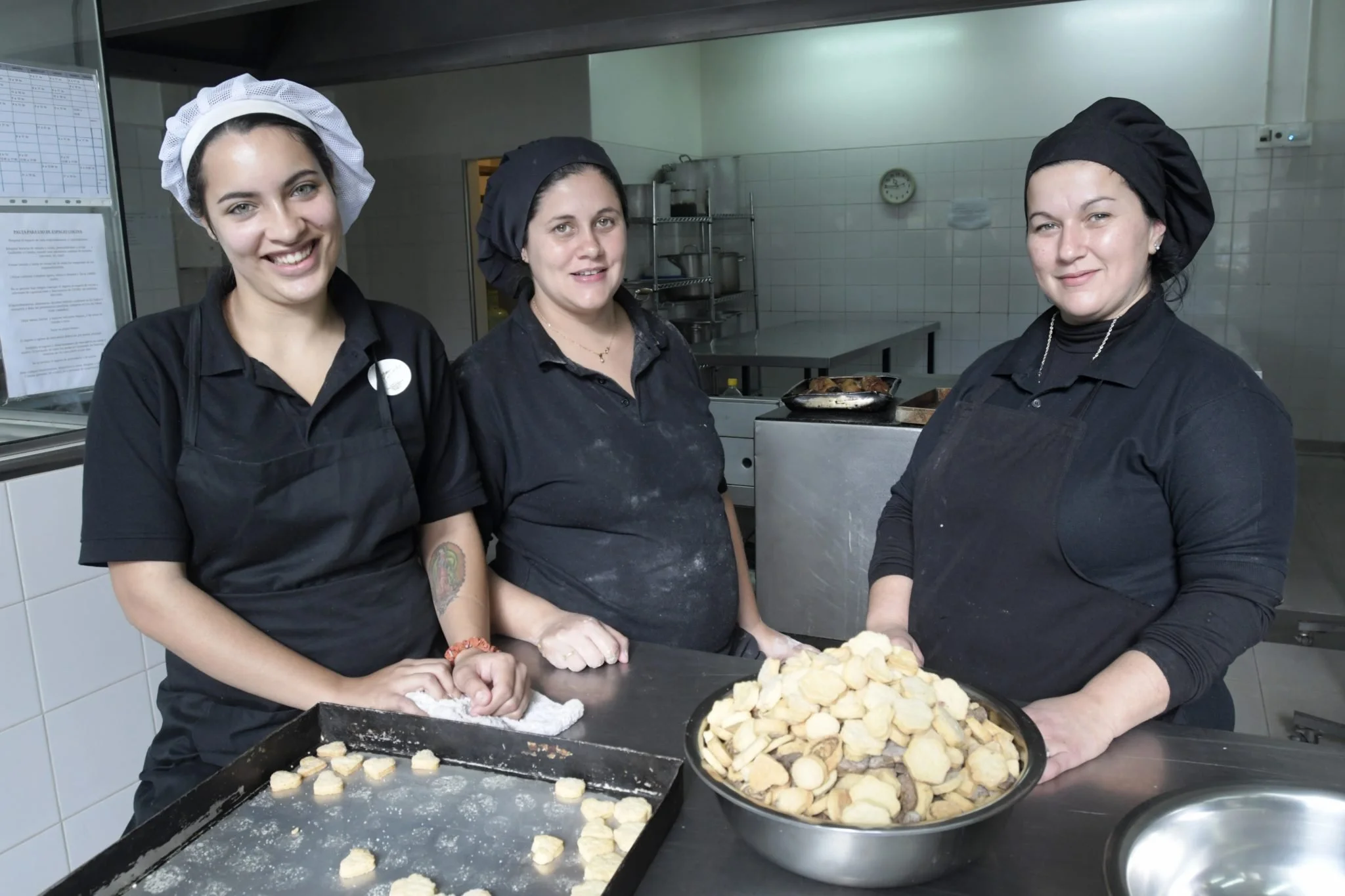 Three women in black uniforms and chef hats in a commercial kitchen, preparing and organizing cut potatoes for cooking or baking.