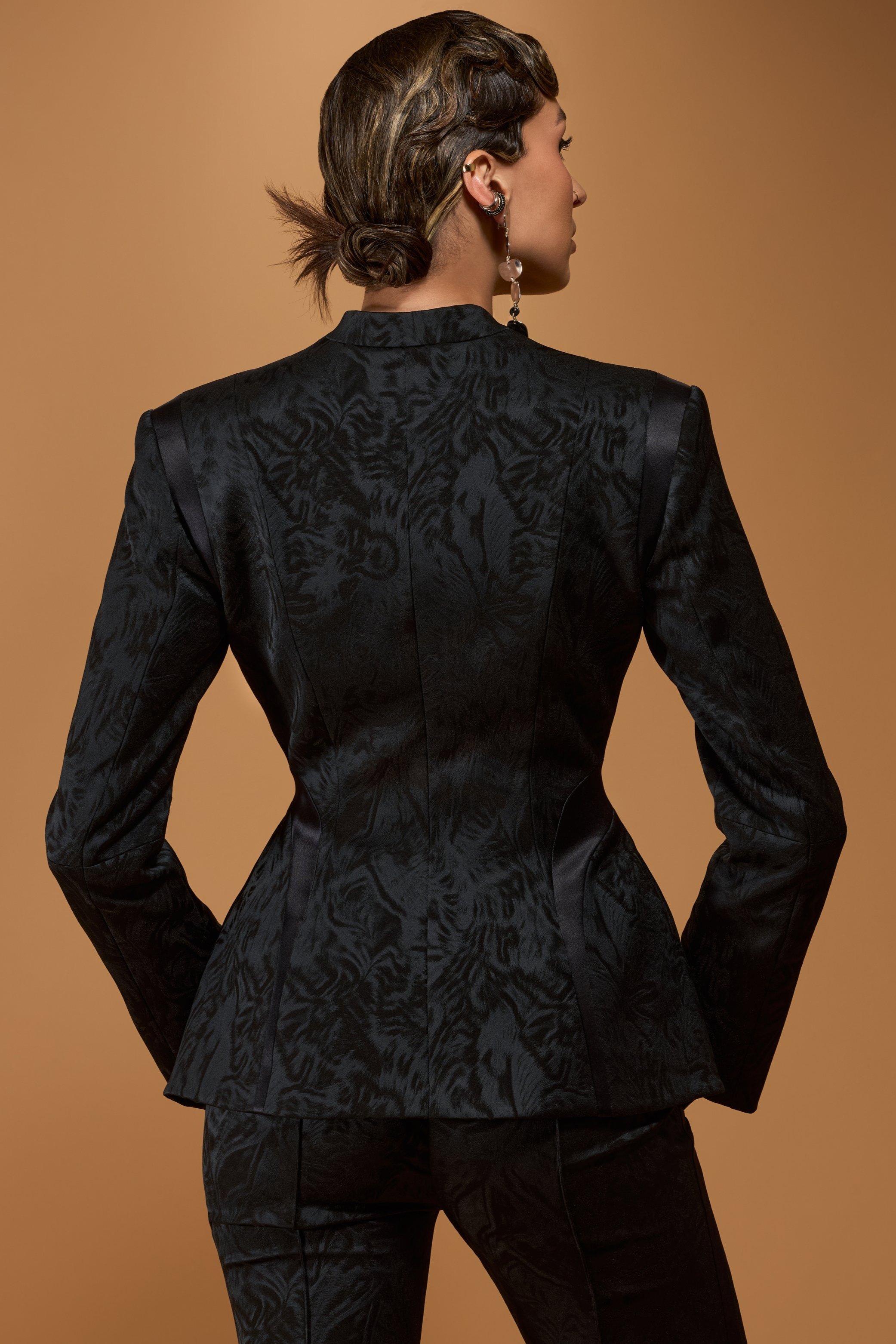 Back view of a woman with styled brown hair wearing a black textured blazer and matching pants, against a tan background.