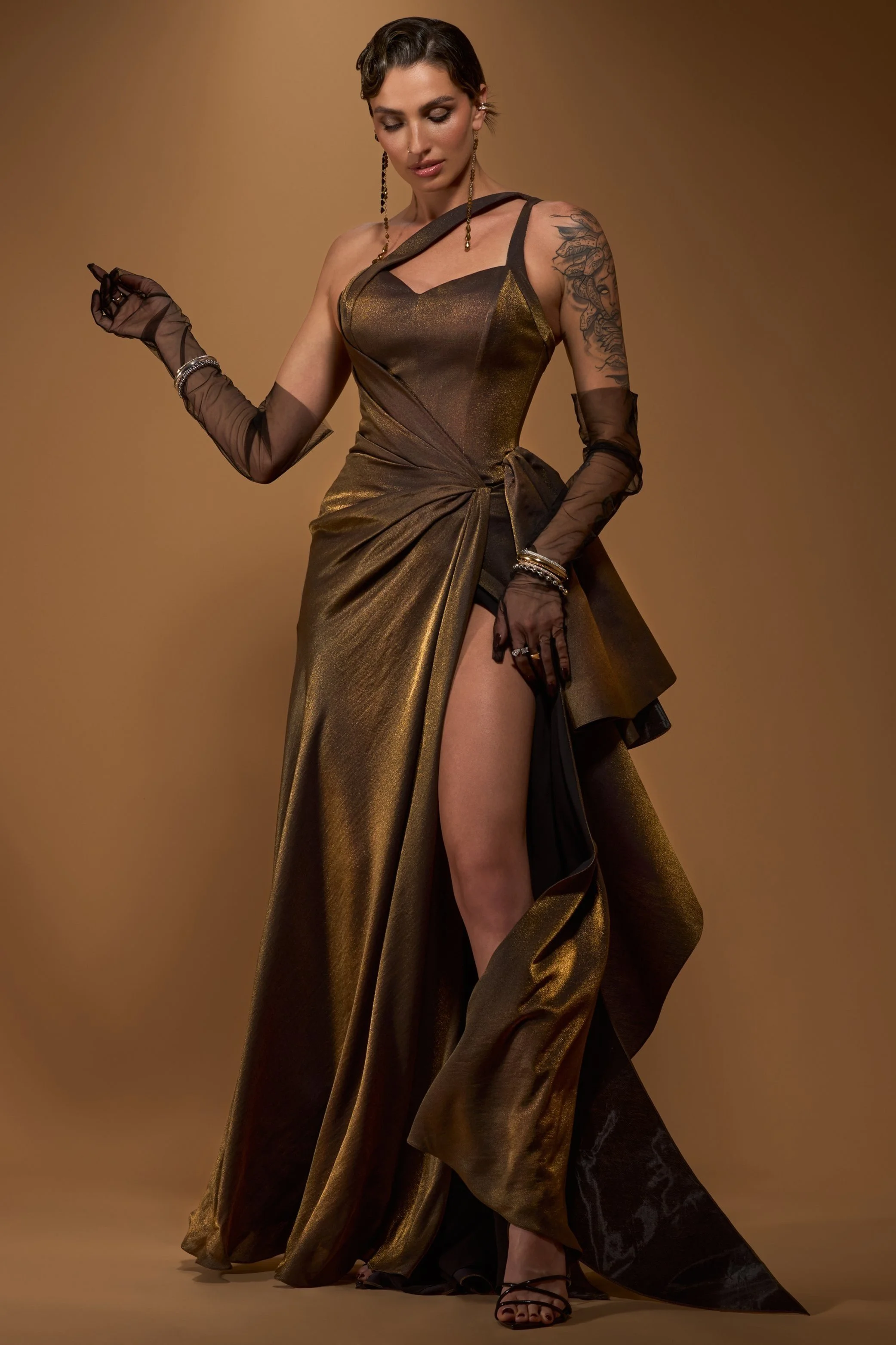 A woman in a gold and black evening gown with high slit, black gloves, and jewelry, posing against a beige background.
