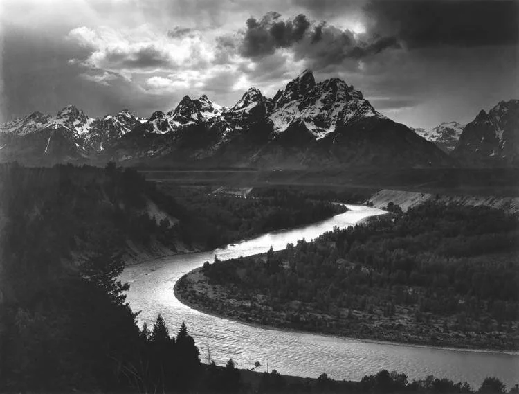 Black and white photo by Ansel Adams of the Grand Tetons behind a silvery Snake River in Jackson Hole, Wyoming with a moody sky.