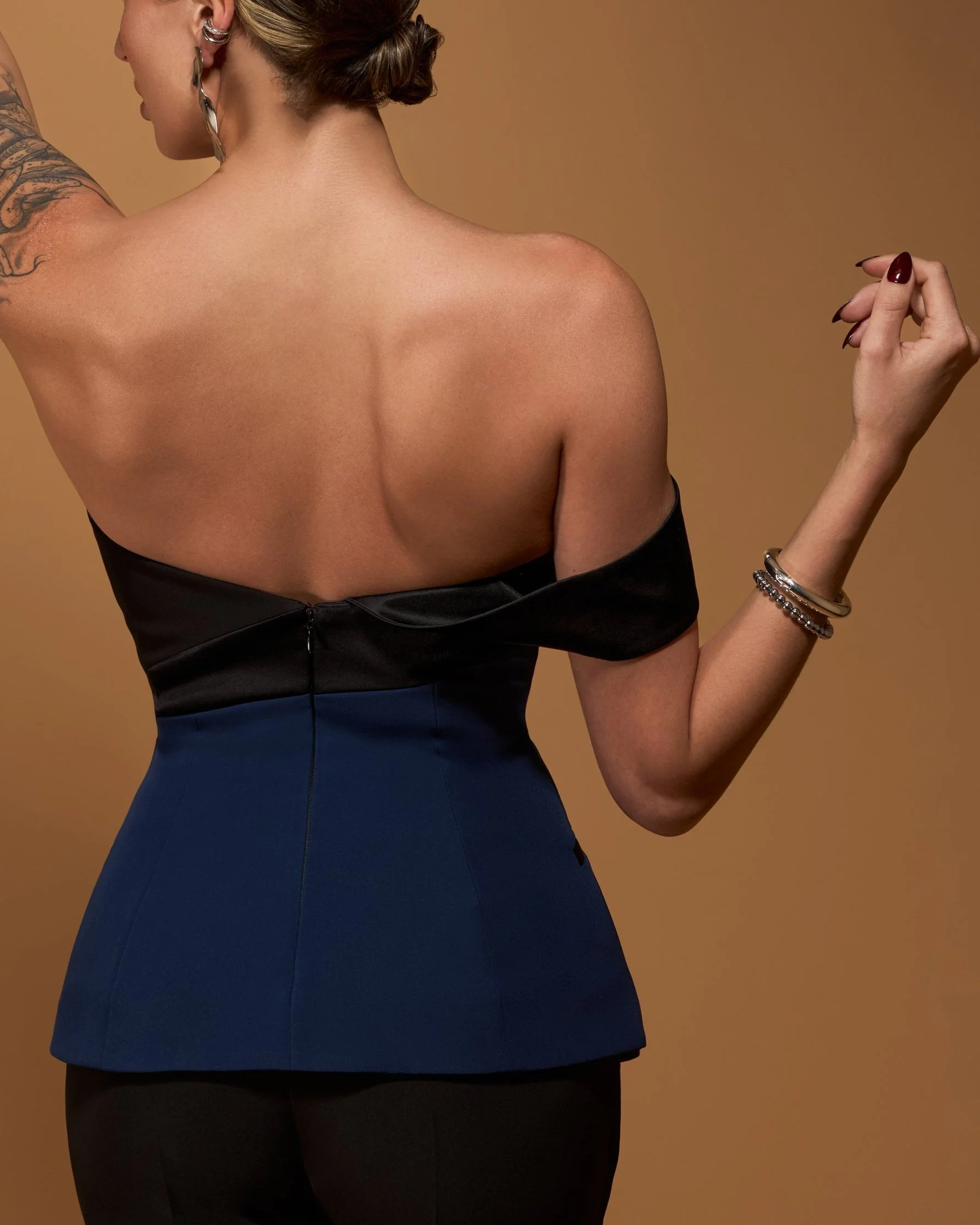 Back view of a woman with modern styled hair, wearing a black and blue off-the-shoulder asymmetrical suiting top, with jewelry on her right wrist, standing against a warm beige background.