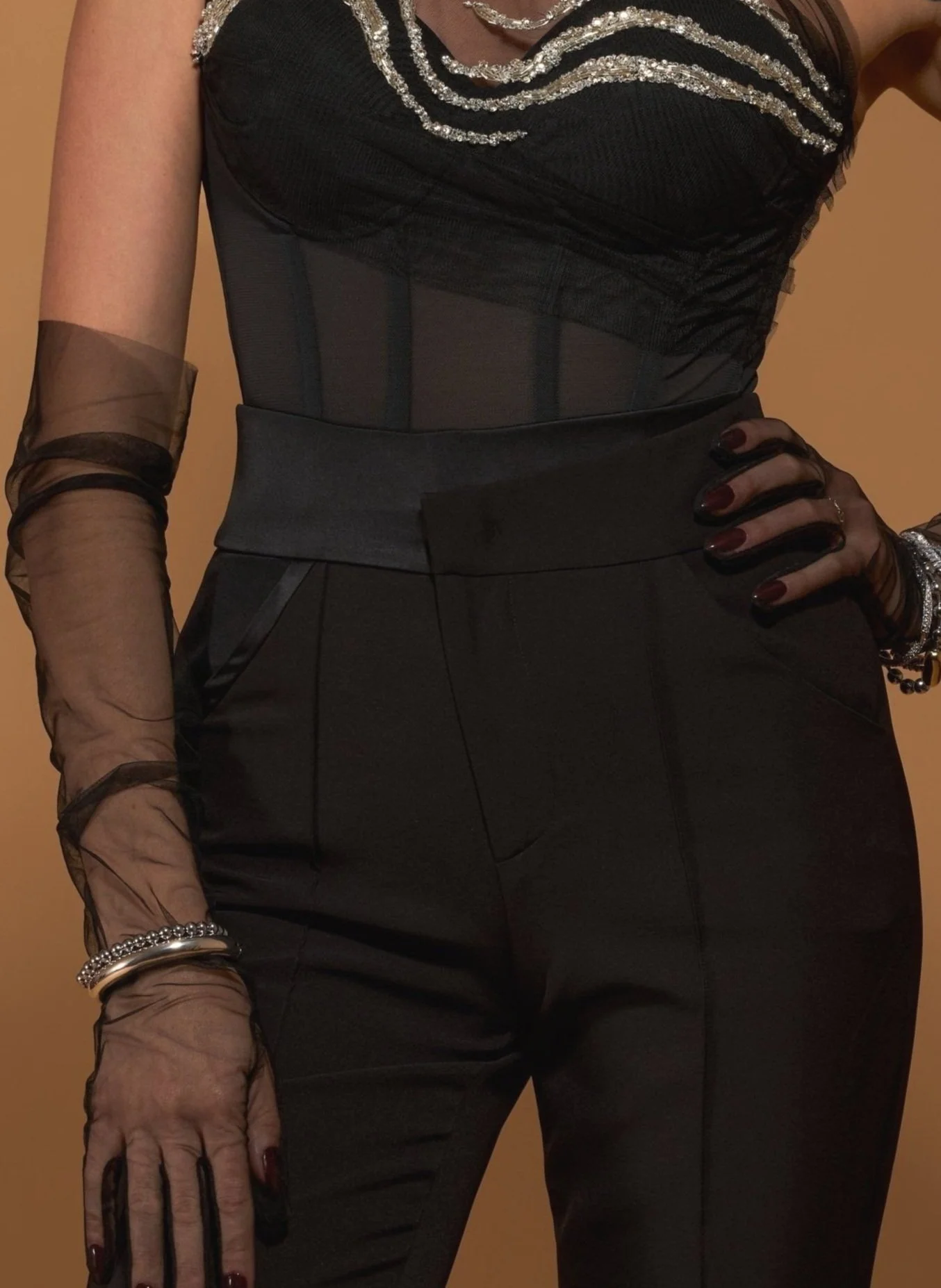 A model wearing a black bustier top with sheer accents beaded with crystal embroidery along the neckline wearing sheer black opera gloves layered with modern silver jewelry, with black suiting pants featuring curved, asymmetrical satin details.