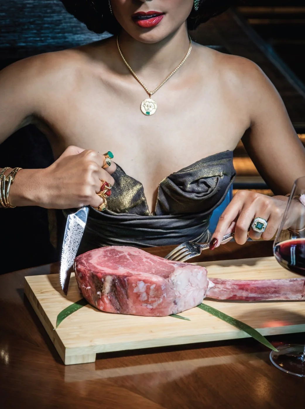 Woman wearing jewelry and a strapless dress preparing to cut a large raw steak on a wooden board, with a glass of red wine nearby.