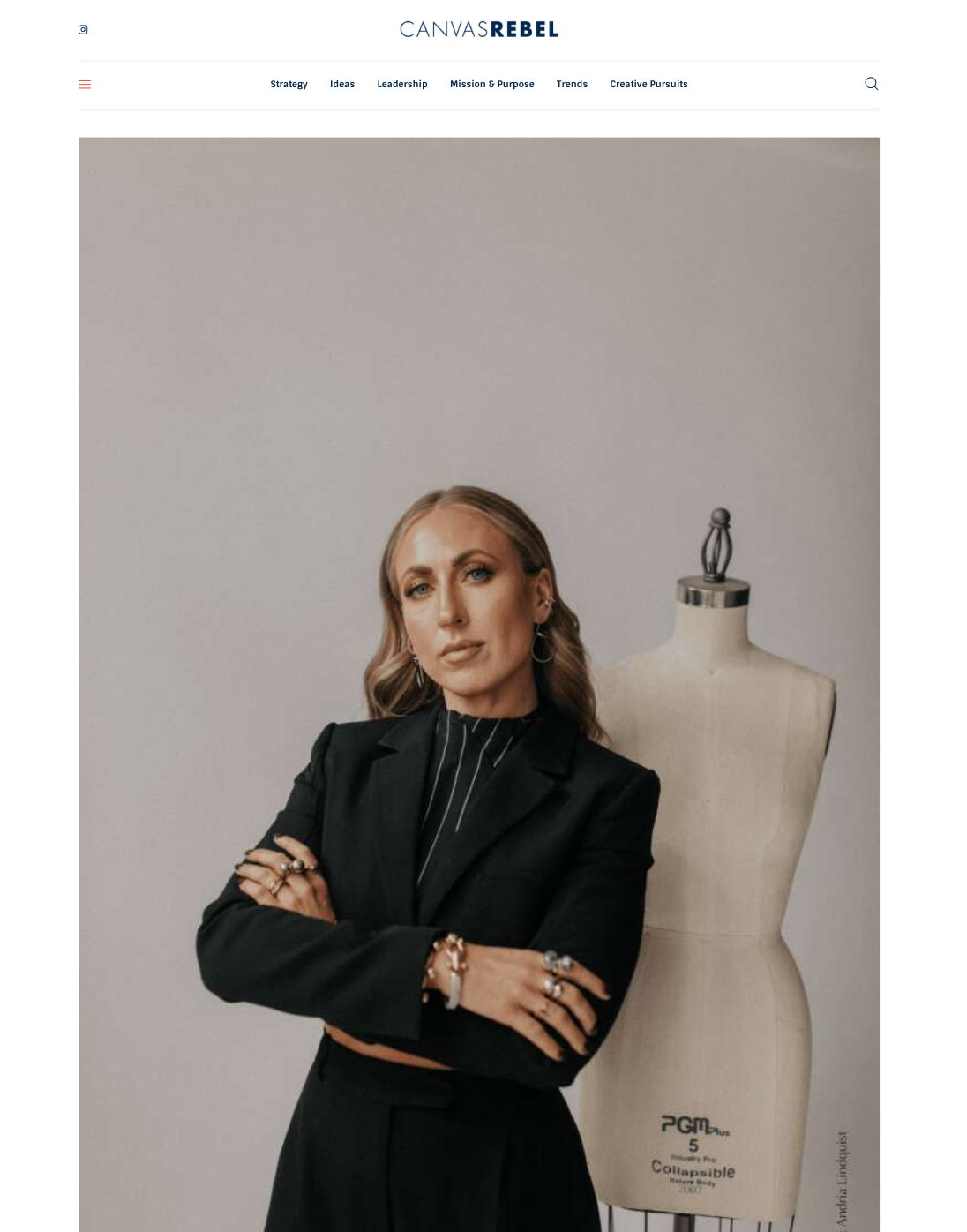 A woman dressed in a black blazer and jewelry, standing with her arms crossed beside a dress form mannequin in a studio setting.