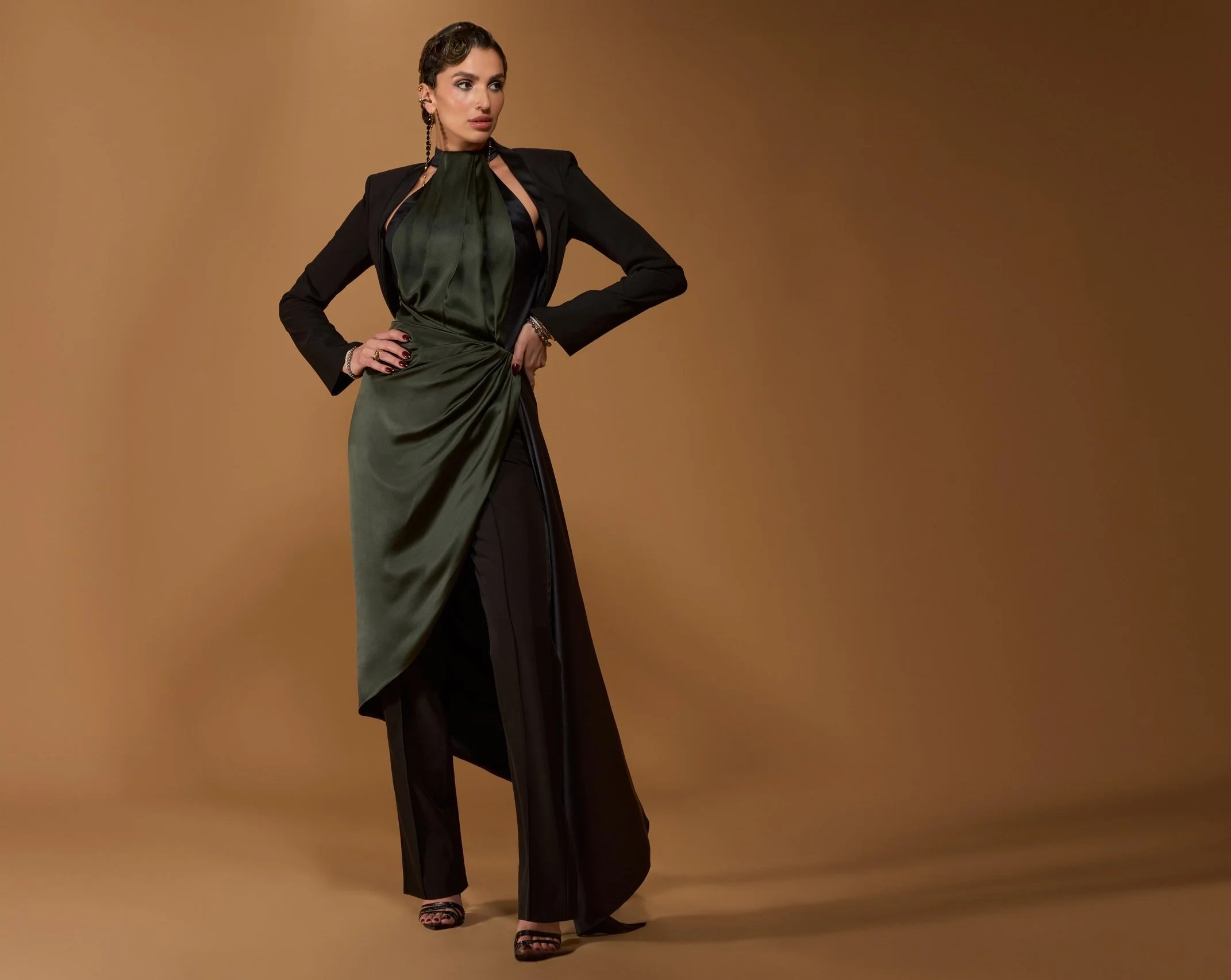 A woman in a fashionable black and green satin evening gown with a wrap detail, pairing it with a black blazer and high heels, standing against a brown background.