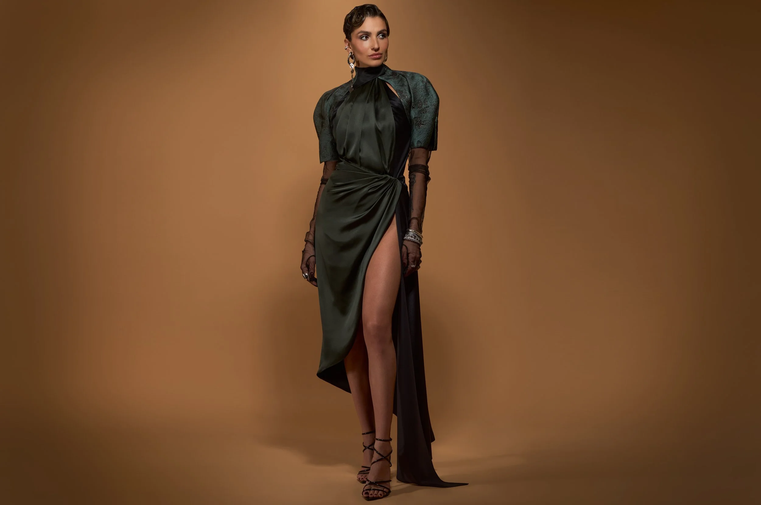 A woman in a black and green evening gown with a high slit, wearing black high heels and sheer gloves, standing against a brown background.