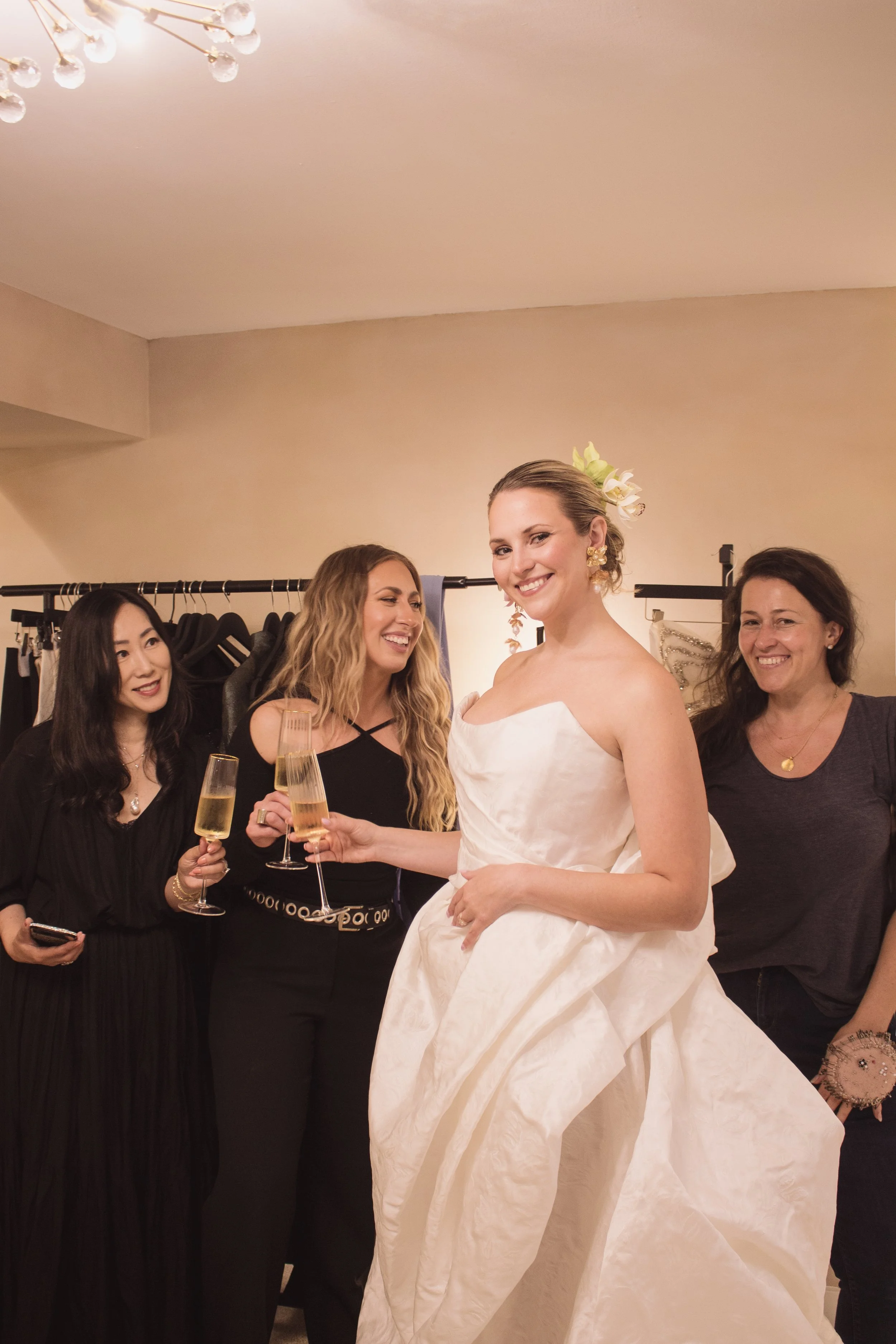 Group of four women celebrating, one in a white wedding dress, holding champagne glasses, smiling, and surrounded by friends in a room with warm lighting.