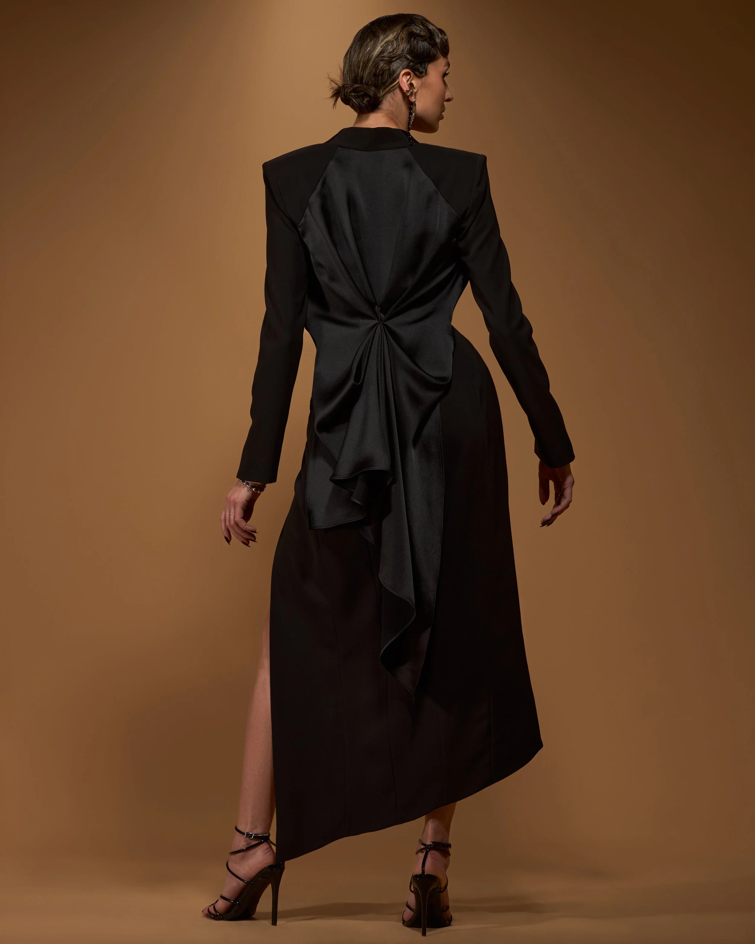 The back of a model wearing a black long sleeve tuxedo jacket with cascading draped black satin back over a black and olive green silk charmeuse asymmetrical angled dress with a thigh high slit.