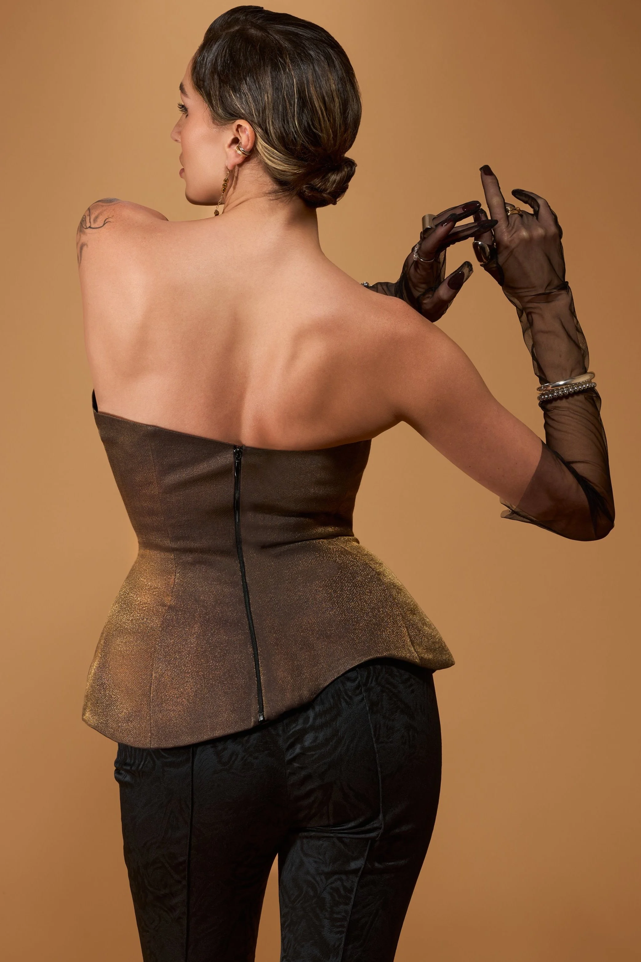 A woman with short, dark hair styled in a low bun, wearing a strapless, shimmery gold-brown top and black patterned pants, with sheer black gloves and jewelry, standing against a plain beige background.