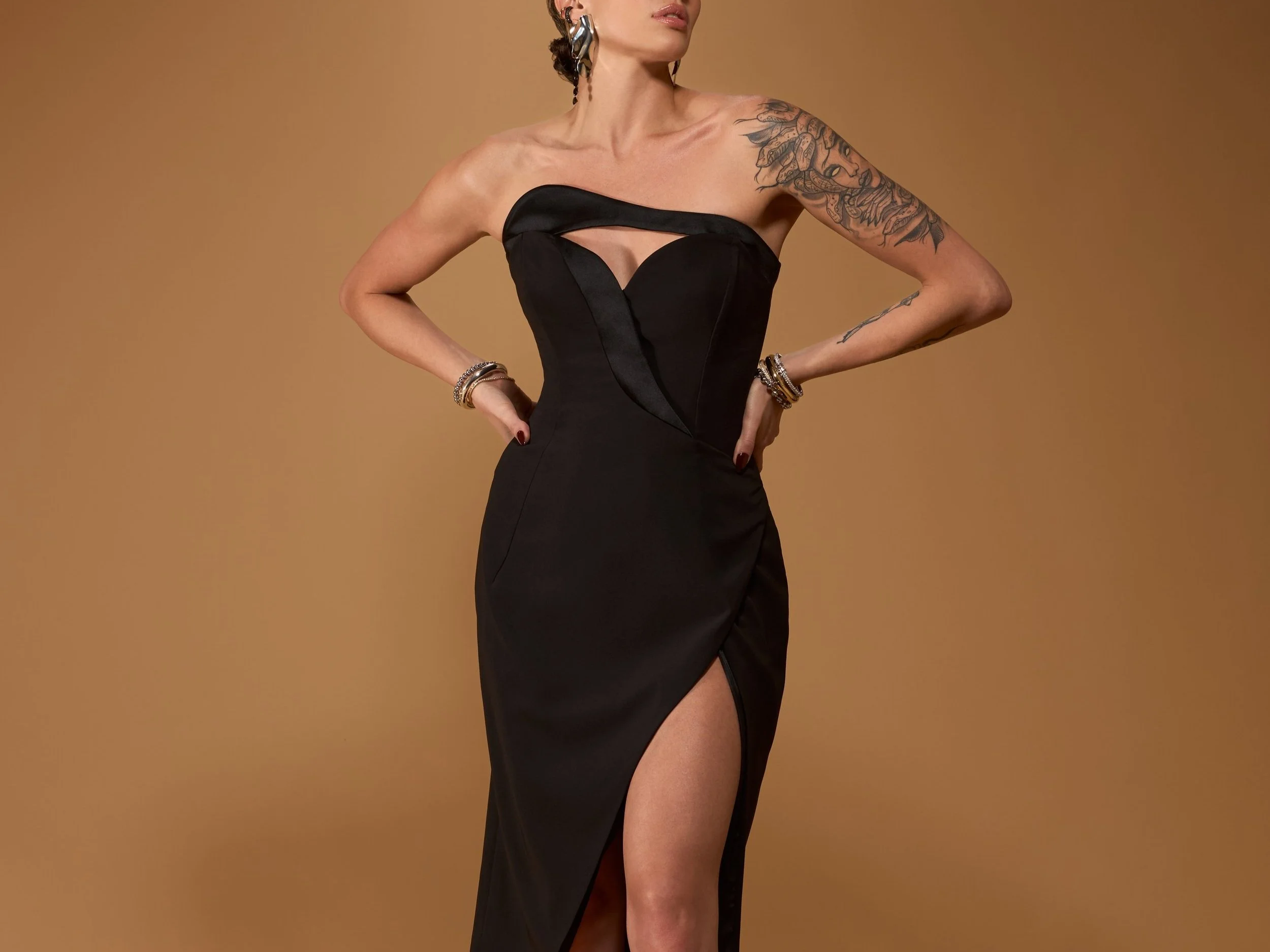 A woman in a black evening gown with a thigh-high slit, posing with her hands on her hips, against a brown background.