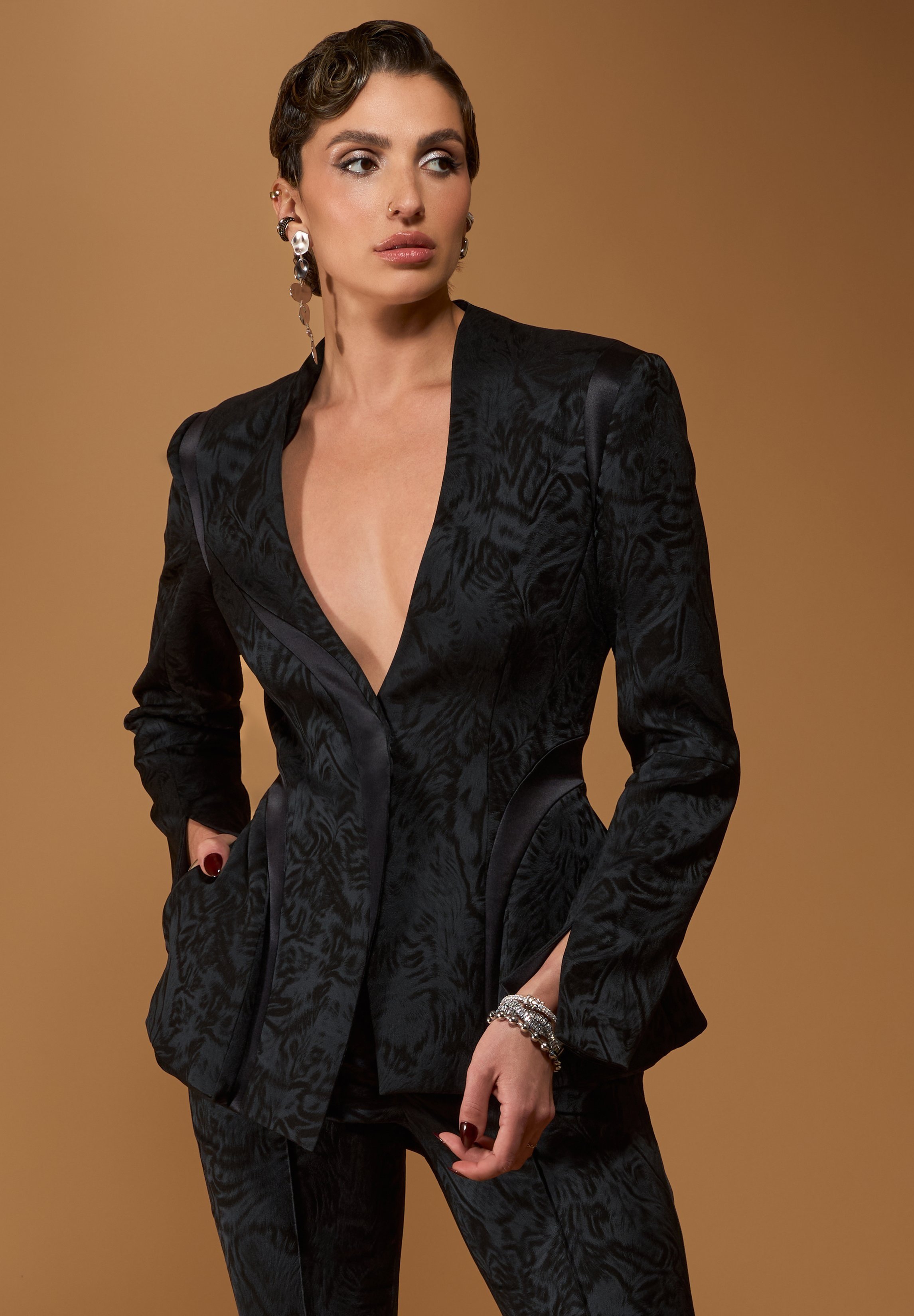 A woman with short wavy hair wearing a black satin textured blazer and matching pants, accessorized with silver earrings, rings, and bracelets, posing against a warm brown background.