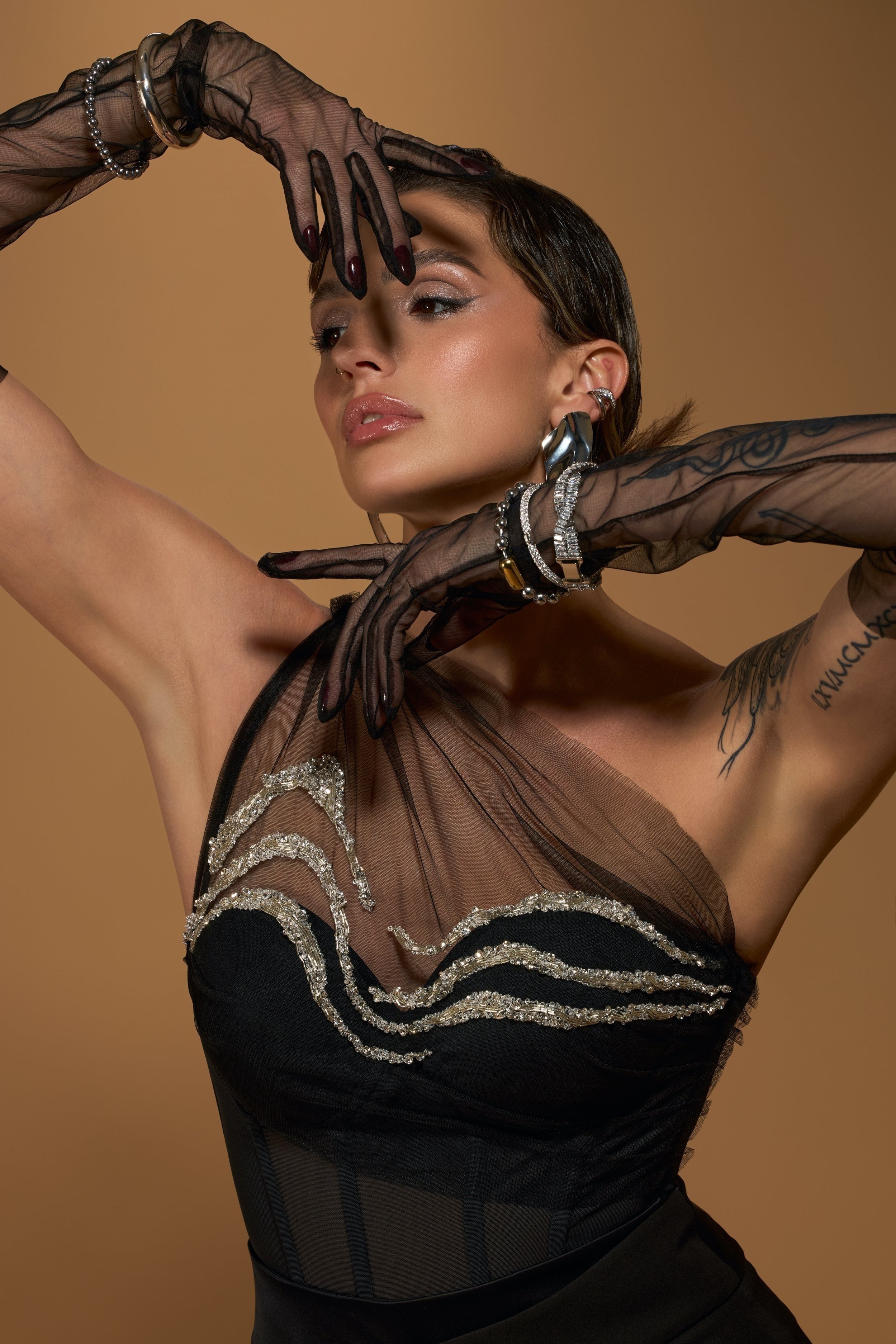 A model wearing a black bustier top with sheer accents beaded with crystal embroidery along the neckline wearing sheer black opera gloves, accessorized with modern silver jewelry, striking an expressive pose with her hands against a beige background.