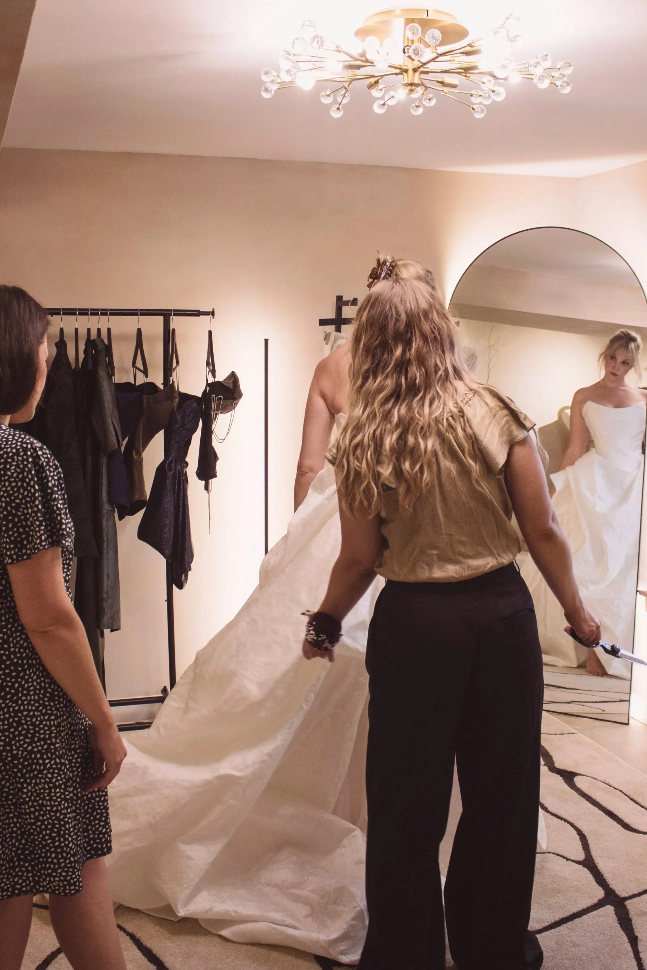 A woman in beige shirt and black pants holding a fabric or garment, assisting a blonde model in a white dress during a fitting or dress fitting session, with a mirror reflecting her.