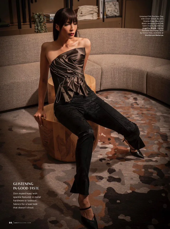 A woman in a strapless satin and silk top with ruffles, black pants, and high heels, sitting on a wooden chair in a modern living room with a curved beige sofa and patterned rug.