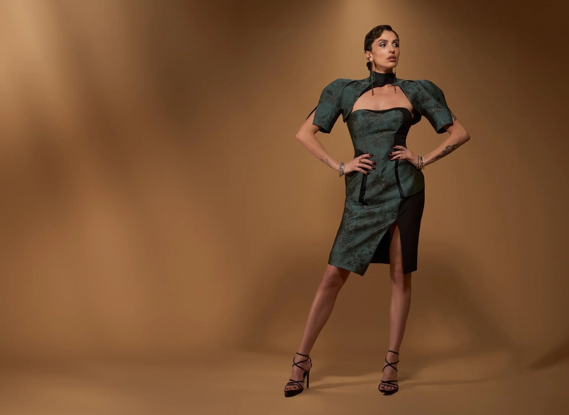 A woman standing confidently with hands on hips, wearing a statement designer dress with a high slit, black heels, and jewelry, against a brown gradient background.