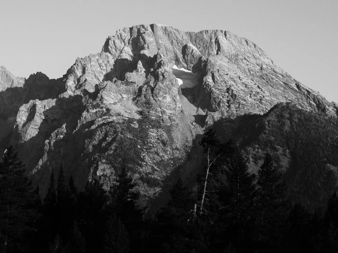 Black and white photo of a rugged mountain with a natural arch formation near the top, surrounded by dense forest at the base.
