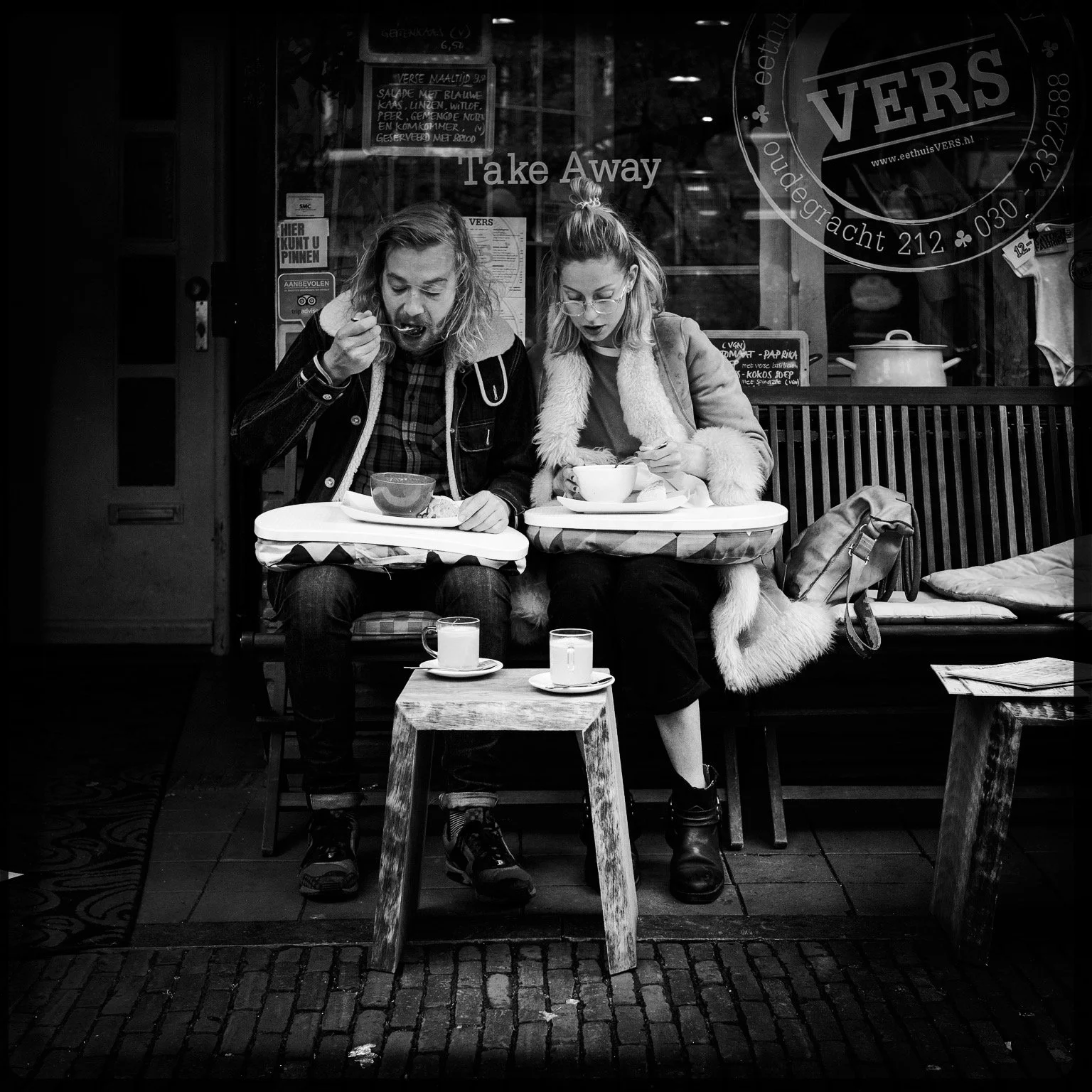 A black and white photo of a man and woman sitting outside a cafe, sharing a meal. The man is eating with a spoon, the woman has a bowl in front of her. There are two cups and a glass of milk on small tables in front of them. They are sitting on a be