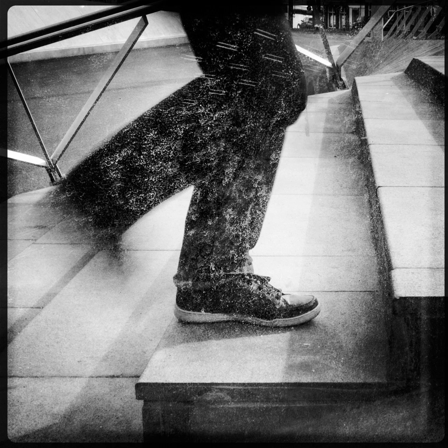 Reflection of a person standing on a sidewalk, wearing dark pants and sneakers, captured through a rain-covered glass window.