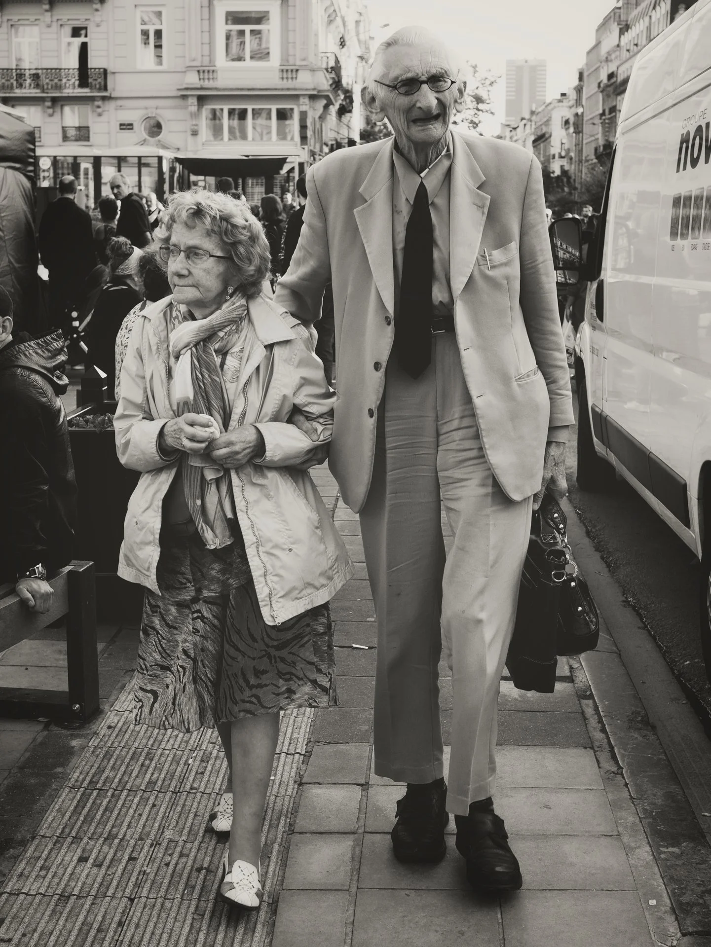 A black-and-white photo of an elderly man and woman holding hands walking on a city sidewalk. The man is wearing a light-colored suit, a dark tie, and glasses, and is looking forward. The woman is wearing a patterned skirt, a light jacket, and a scar
