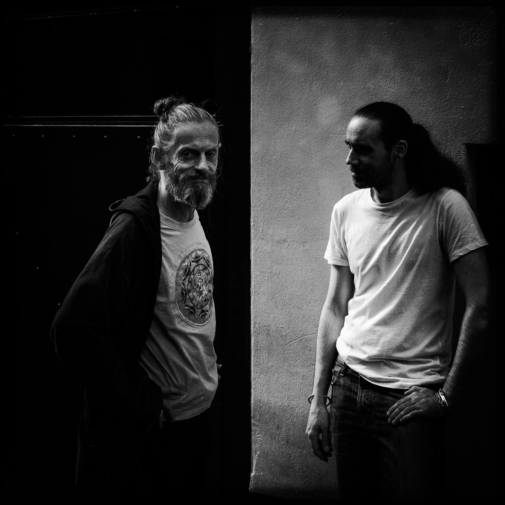 Two men standing in front of a wall, one with a beard and long hair tied back, wearing a T-shirt featuring a circular design, and the other with long hair and a mustache, wearing a plain T-shirt. The photograph is in black and white.