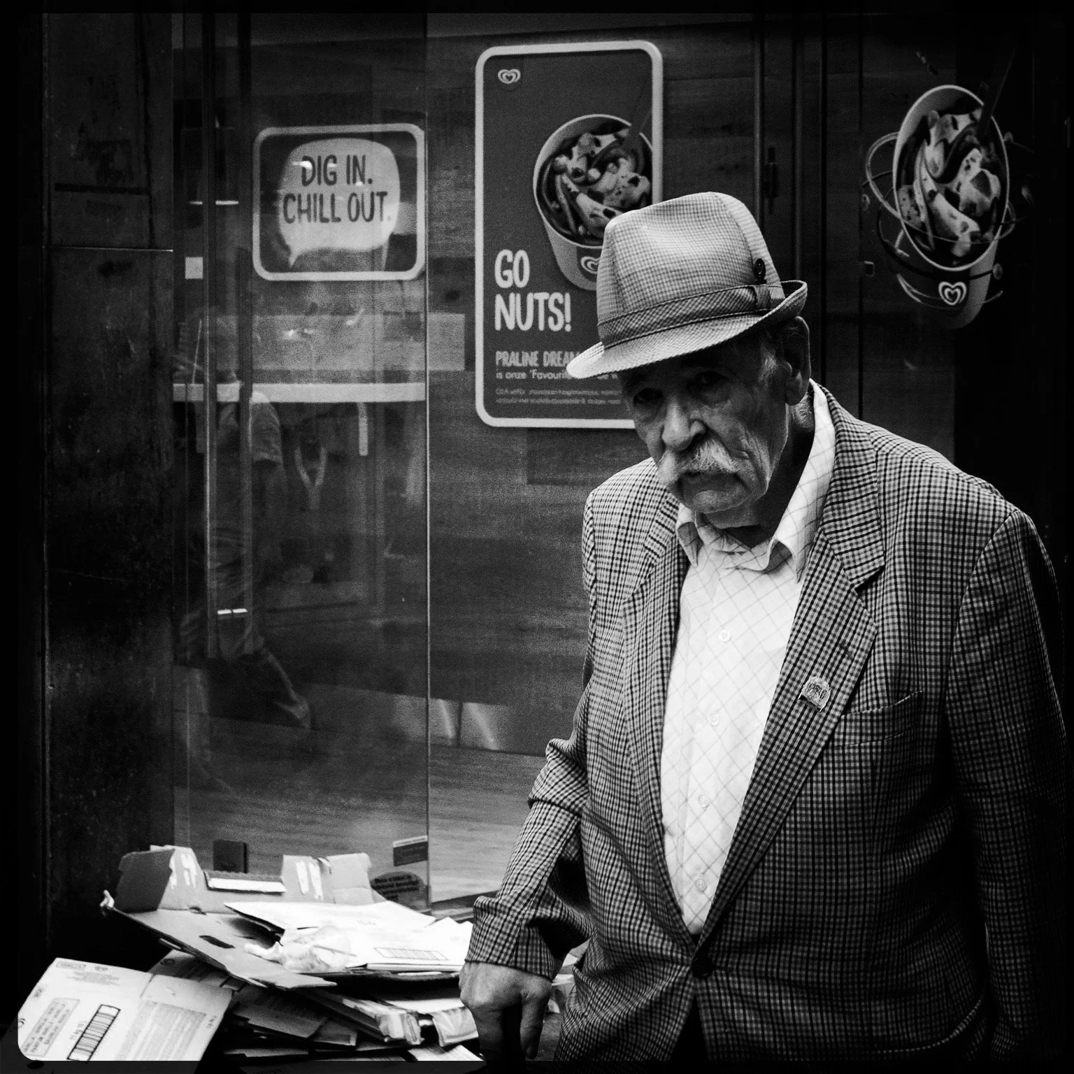 An elderly man with a mustache wearing a checked blazer and a hat, standing in front of a glass door with posters. One poster says "Dig In. Chill Out" and another says "Go Nuts!" with images of ice cream.