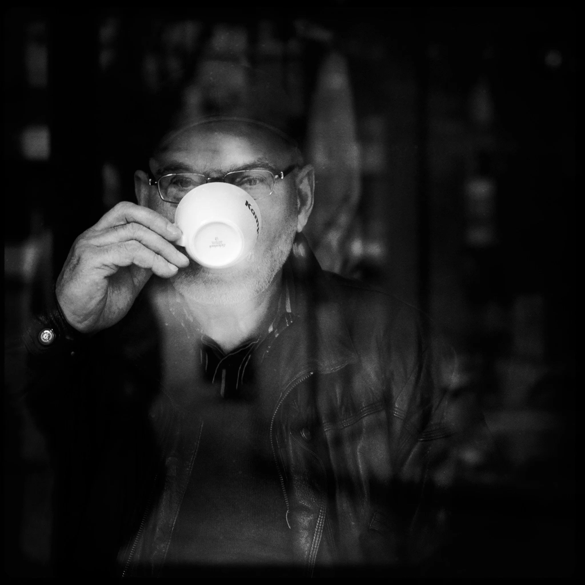 A man with glasses and a leather jacket drinking coffee, photographed through a window, in black and white.