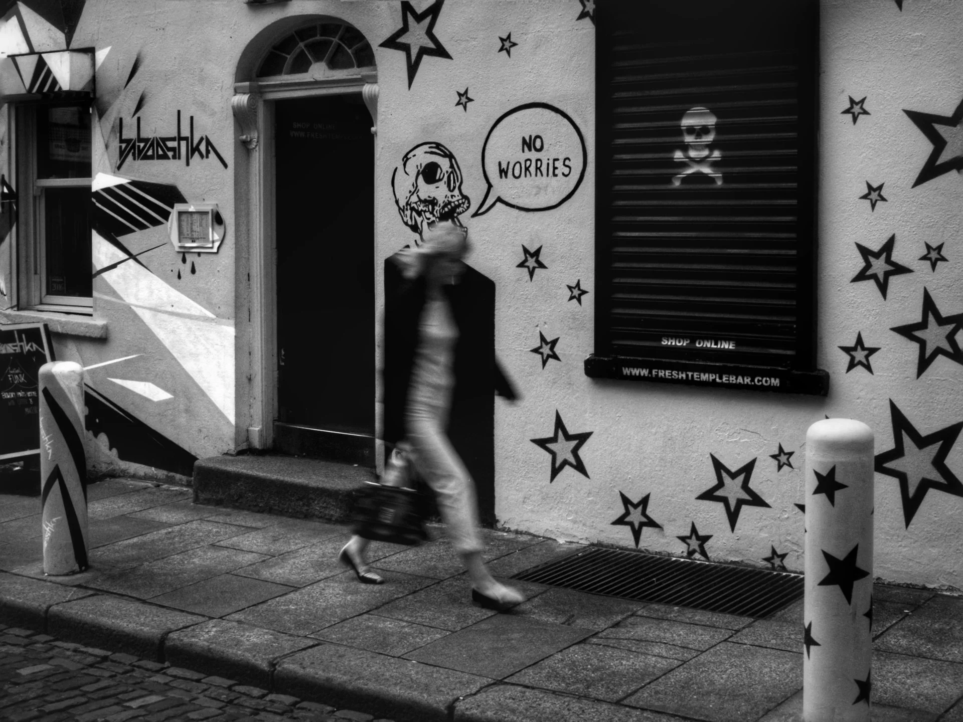 A woman walking past a graffiti-covered building with cartoon skulls and star decorations, a speech bubble that says 'No Worries', cobblestone sidewalk, bollards with star patterns, and a sign for a bar called 'Freshtemplebar'.