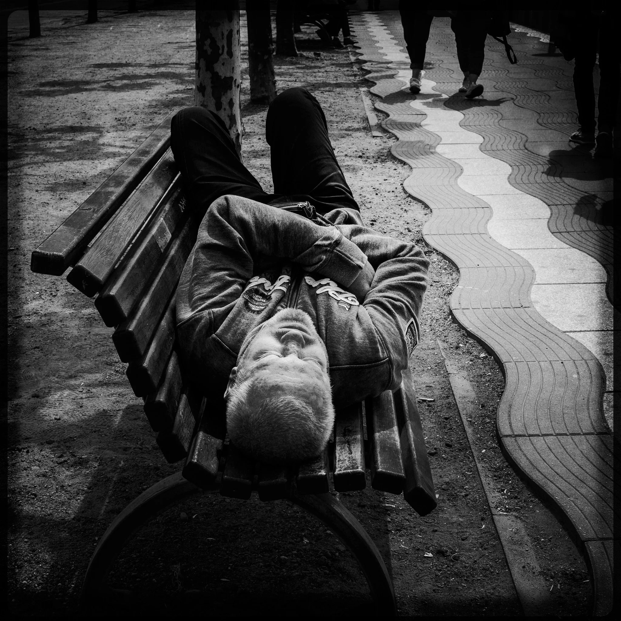 A man lying down on a park bench with his arms crossed over his chest, with a paved sidewalk and several walking people in the background.