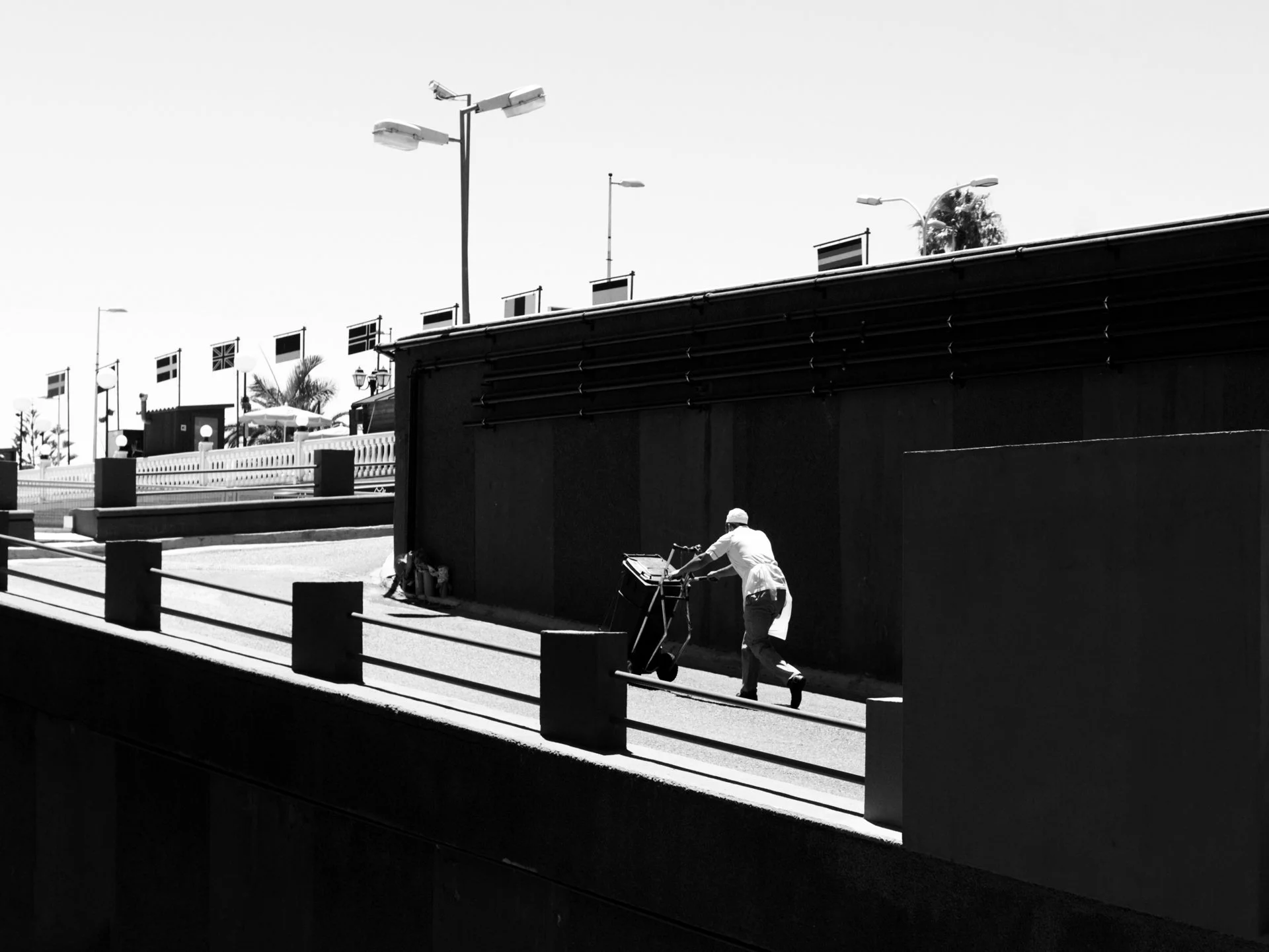 A person pushing a shopping cart along a sidewalk near a dark wall with a bright sky overhead.