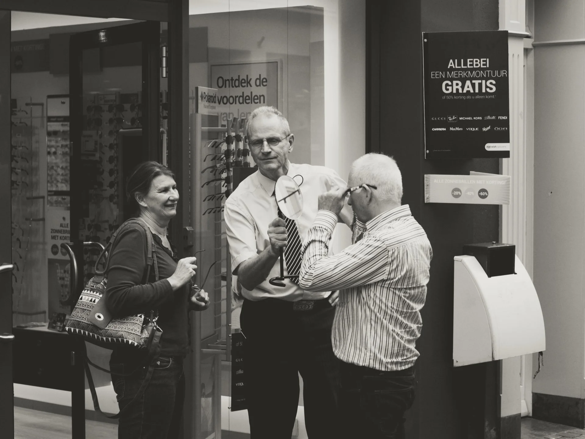 Three people, two men and one woman, are standing outside a store entrance. One man with glasses and a striped shirt is trying on a pair of glasses to the man who is not wearing glasses. The woman is smiling and holding a bag. There are signs in Dutc