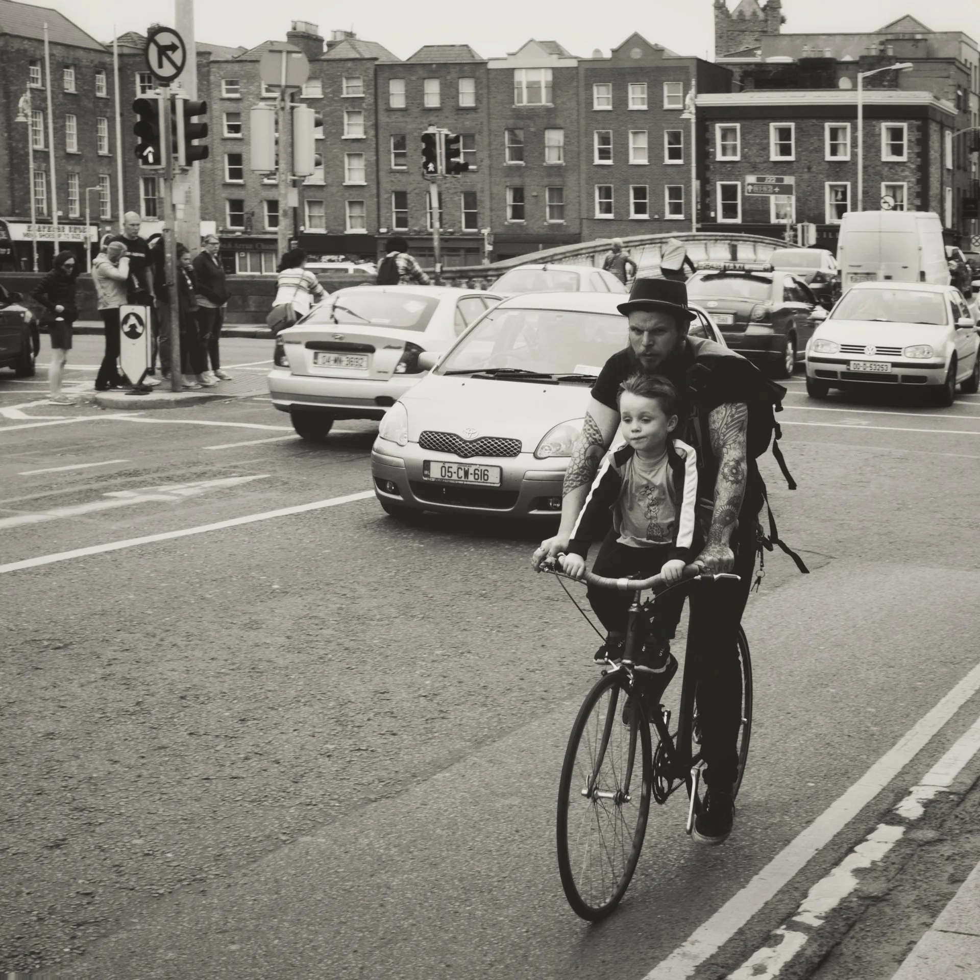 A man and a young boy riding a bicycle together on a city street, with cars parked and buildings in the background.