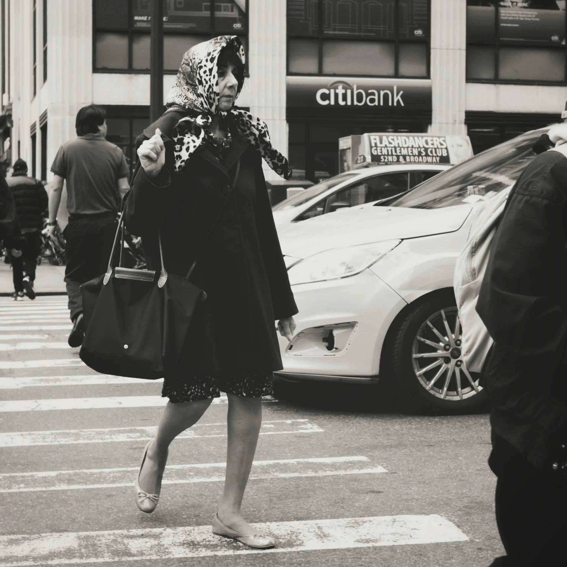 Black and white photo of a woman crossing a street, wearing a leopard print scarf on her head and a dark coat, with a handbag in her left hand. Cars and buildings are visible in the background, including a Citibank sign and billboard for Flashdancers