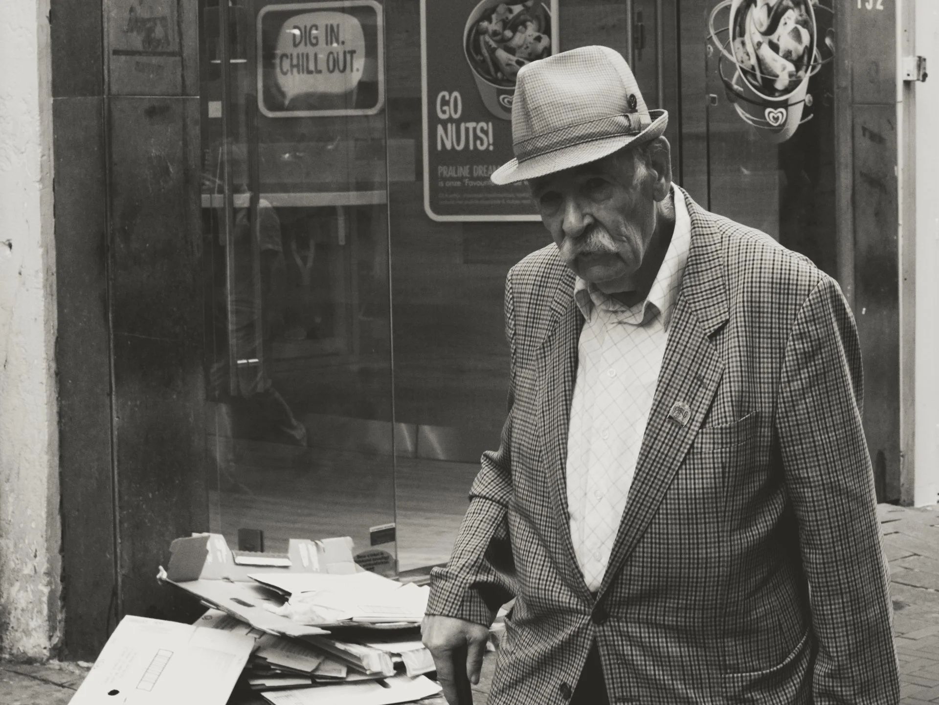 An elderly man with a mustache wearing a checkered blazer, a light-colored shirt, and a hat, standing outdoors in front of a glass storefront. There are cardboard boxes and papers on the ground in front of him, and a sign inside the store reads 'DIG 