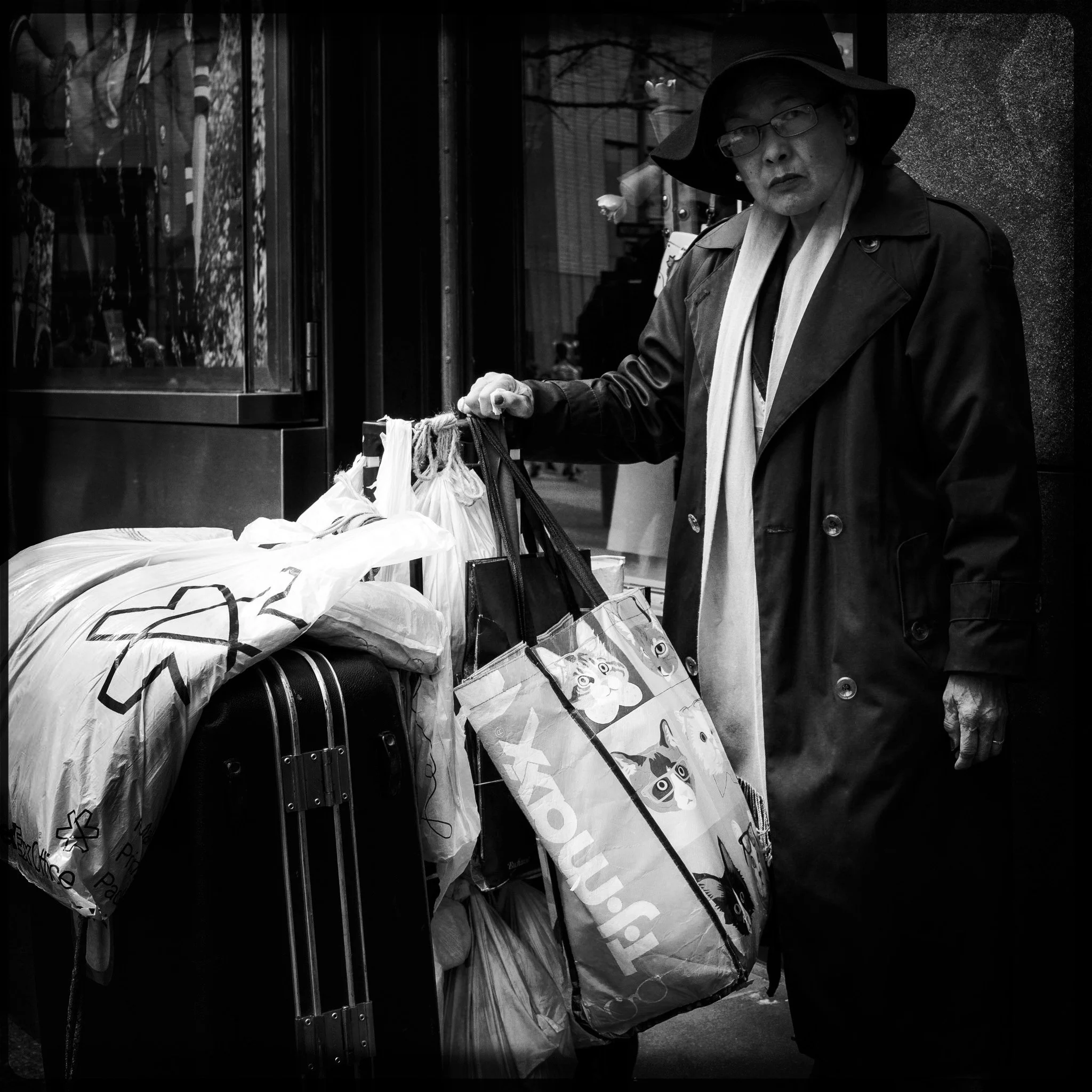 A woman in a coat and large hat looks at the camera while standing outdoors, holding shopping bags and near a suitcase, with various bags and packages around her.