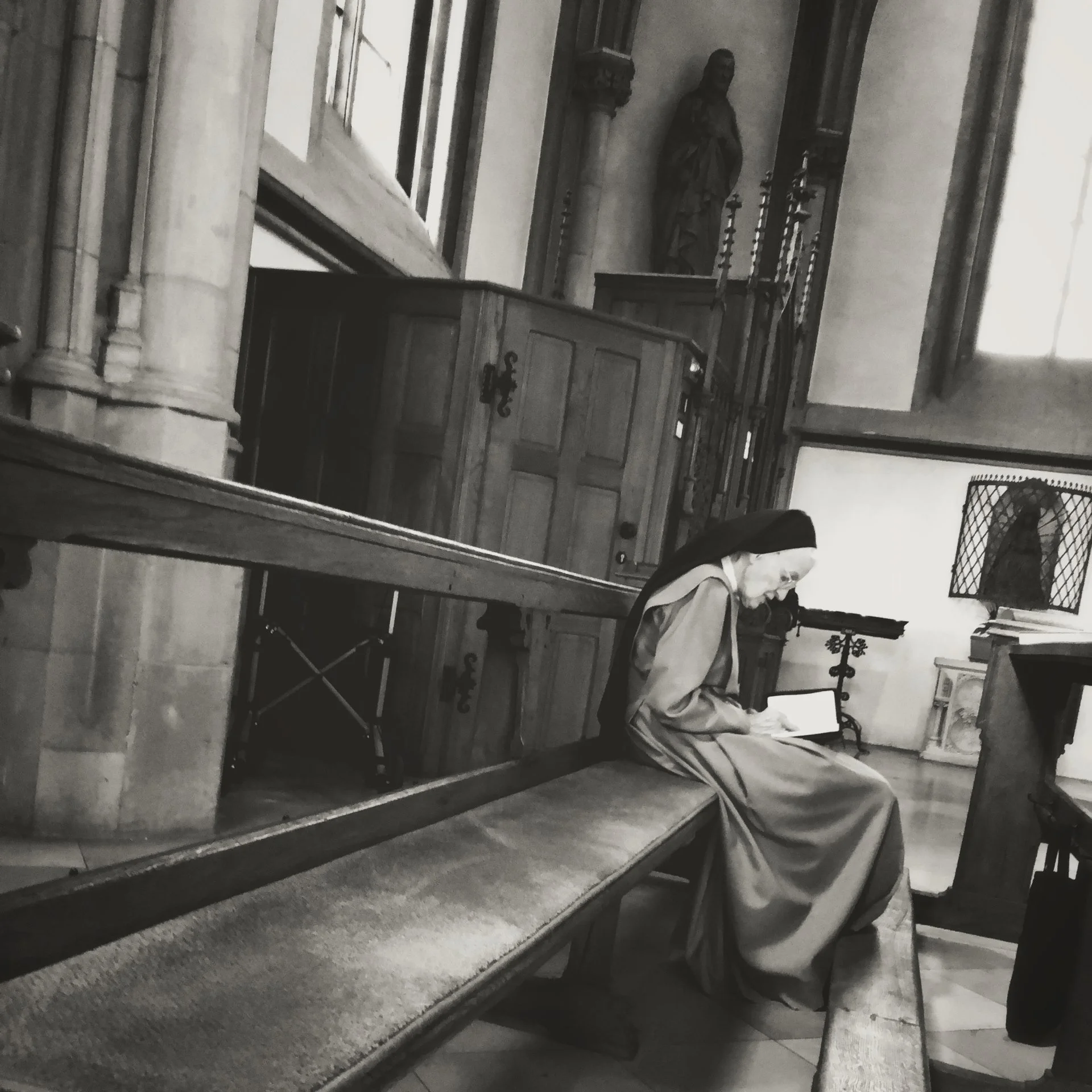A woman sitting on a pew in a church, reading a book, with religious statues and candles in the background, inside a historic building.