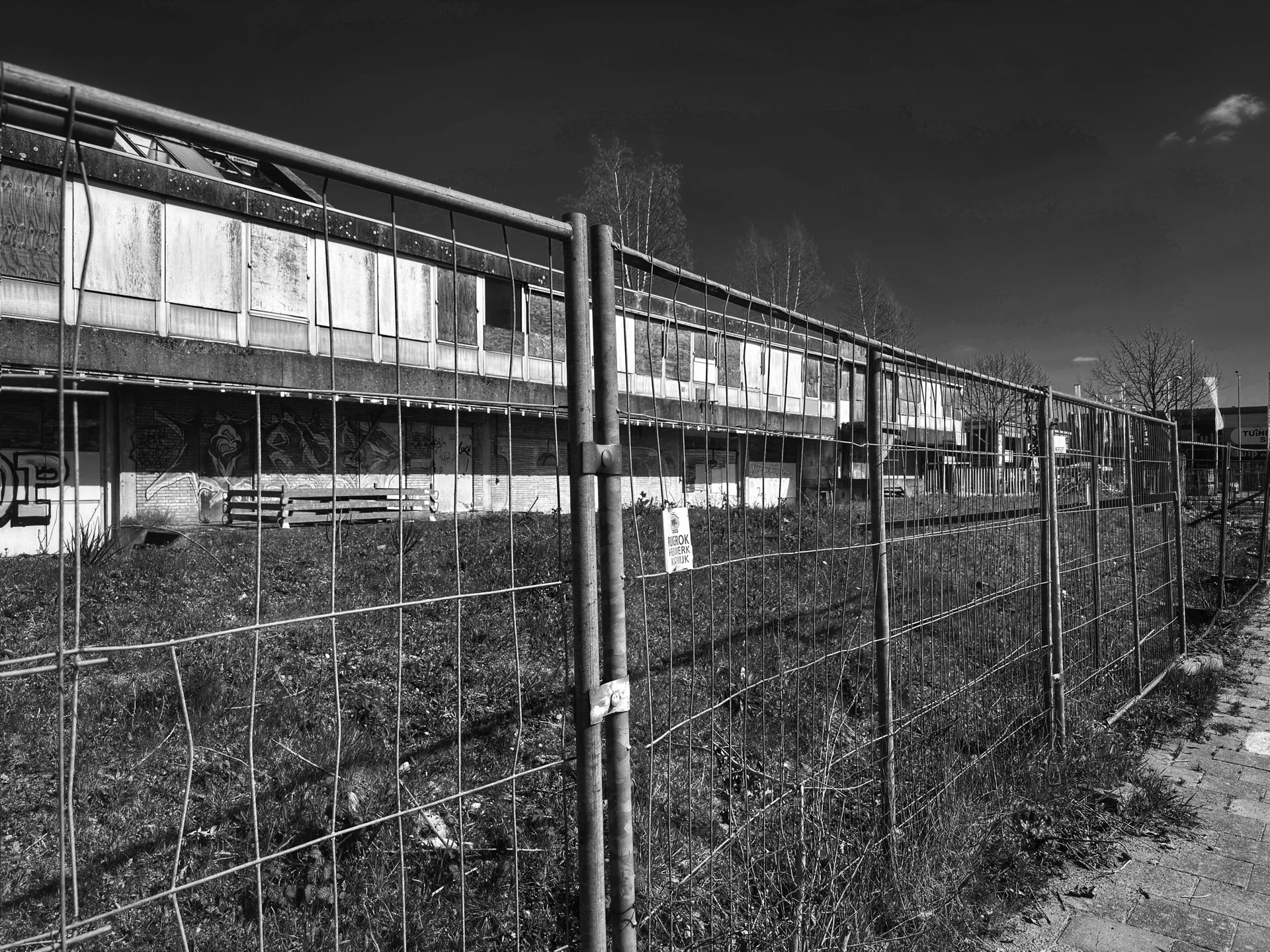 An abandoned building behind a chain-link fence, with graffiti on the walls and leafless trees in the background, under a partly cloudy sky.