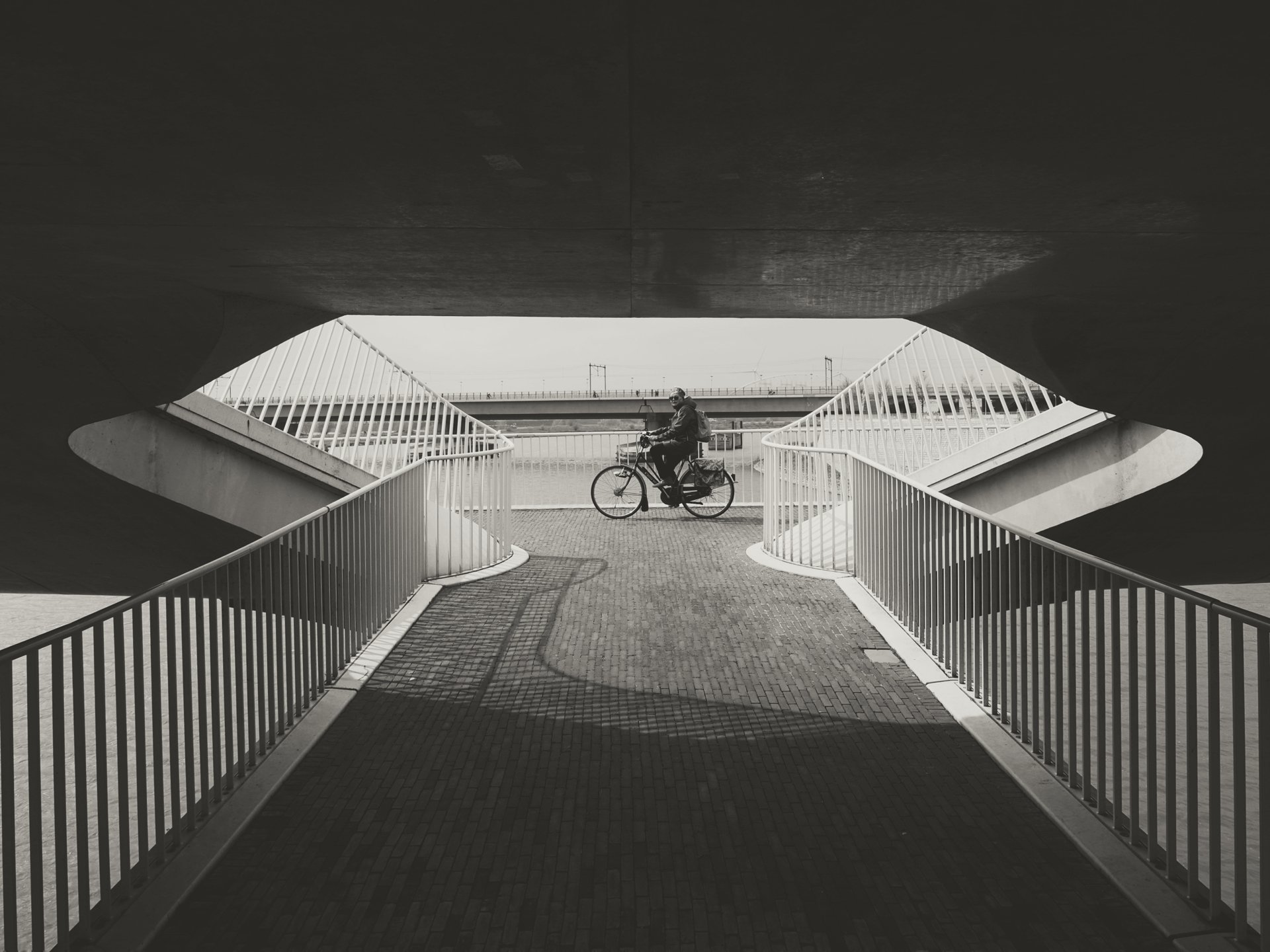 A man riding a bicycle under a dark overpass with a pedestrian bridge in the background.