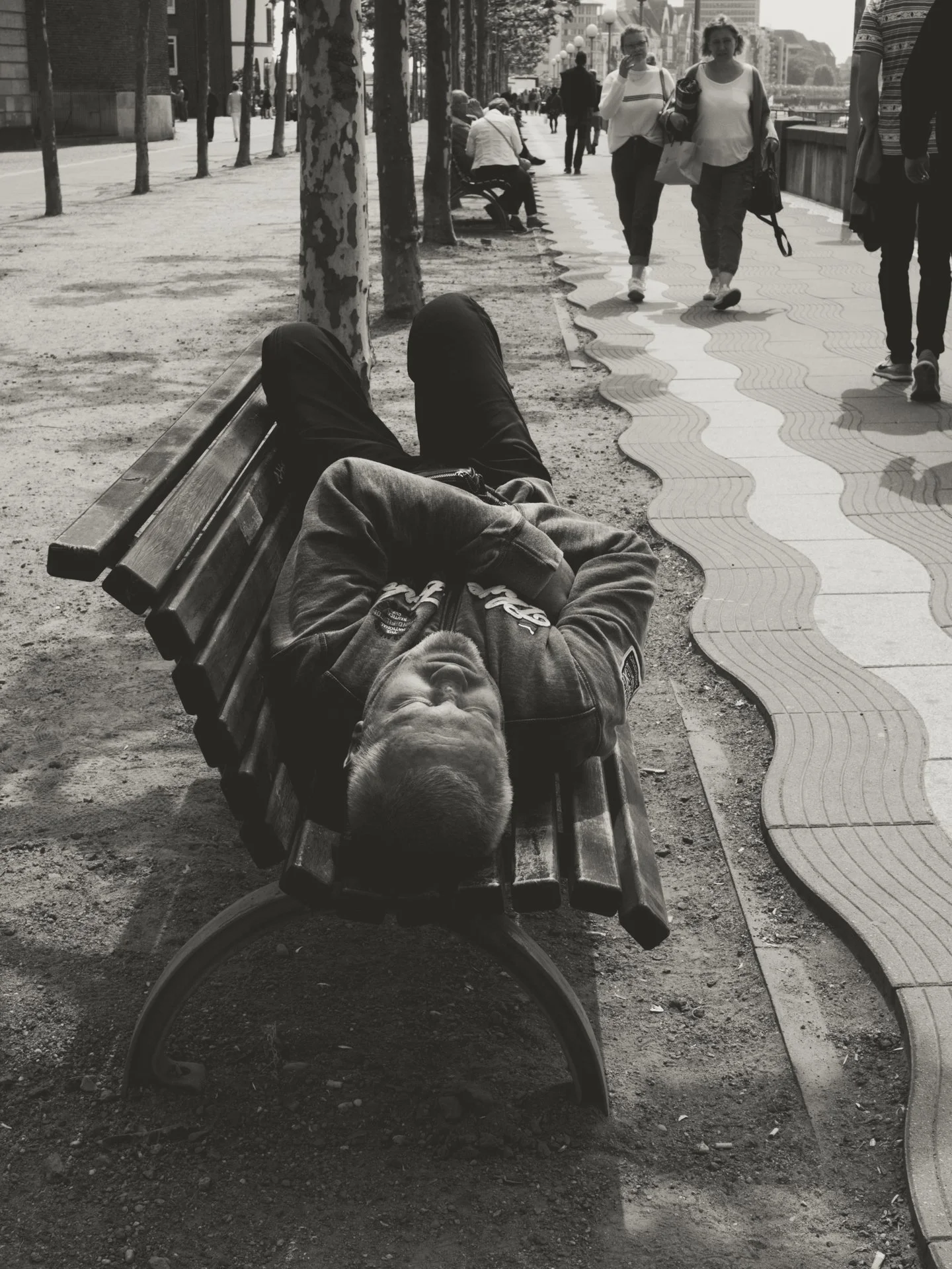 A person lying on a park bench with legs crossed, wearing dark pants and a jacket, with their head resting on the bench's armrest, facing upwards. In the background, a busy city sidewalk with several pedestrians walking, some sitting on benches, and 