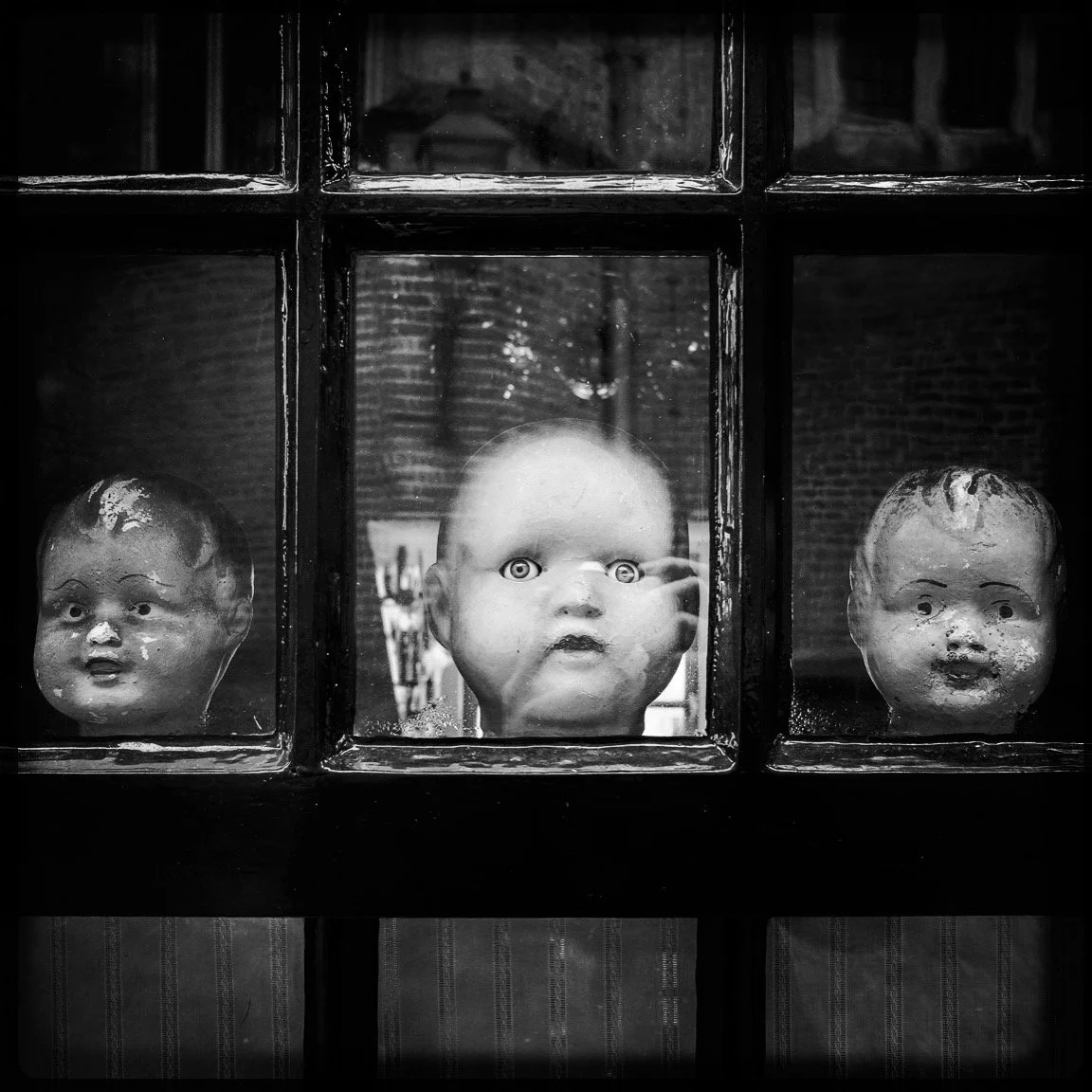 Three old, weathered doll heads displayed behind a glass window, with the central head featuring piercing eyes and a serious expression.