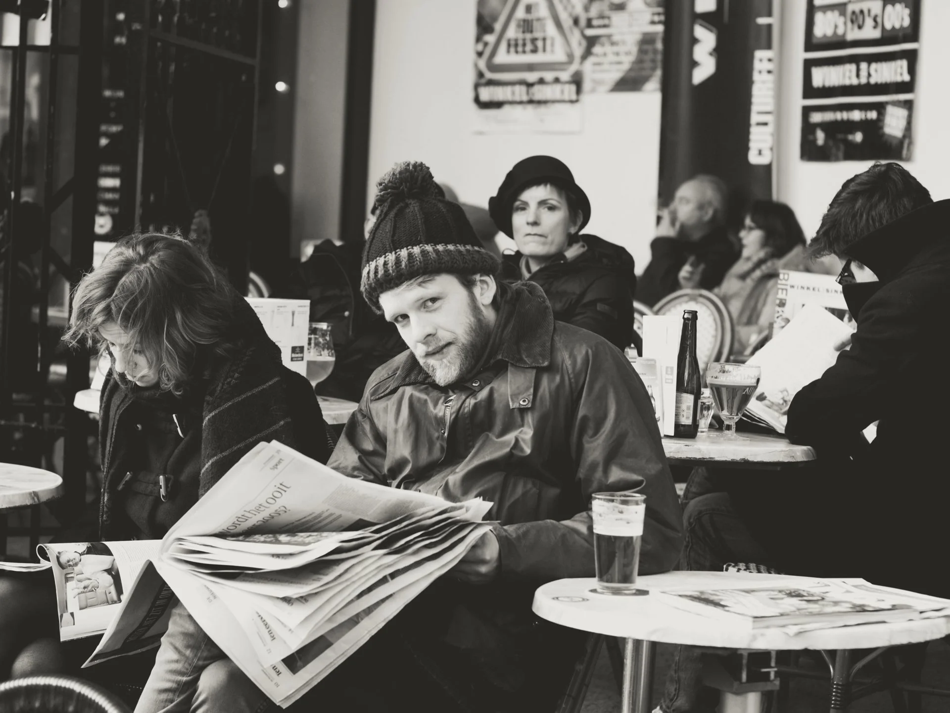A group of people sitting at a table in a cafe or restaurant, engaging in various activities like reading newspapers, drinking beers, and chatting. The background shows posters and signs on the wall, all in black and white.