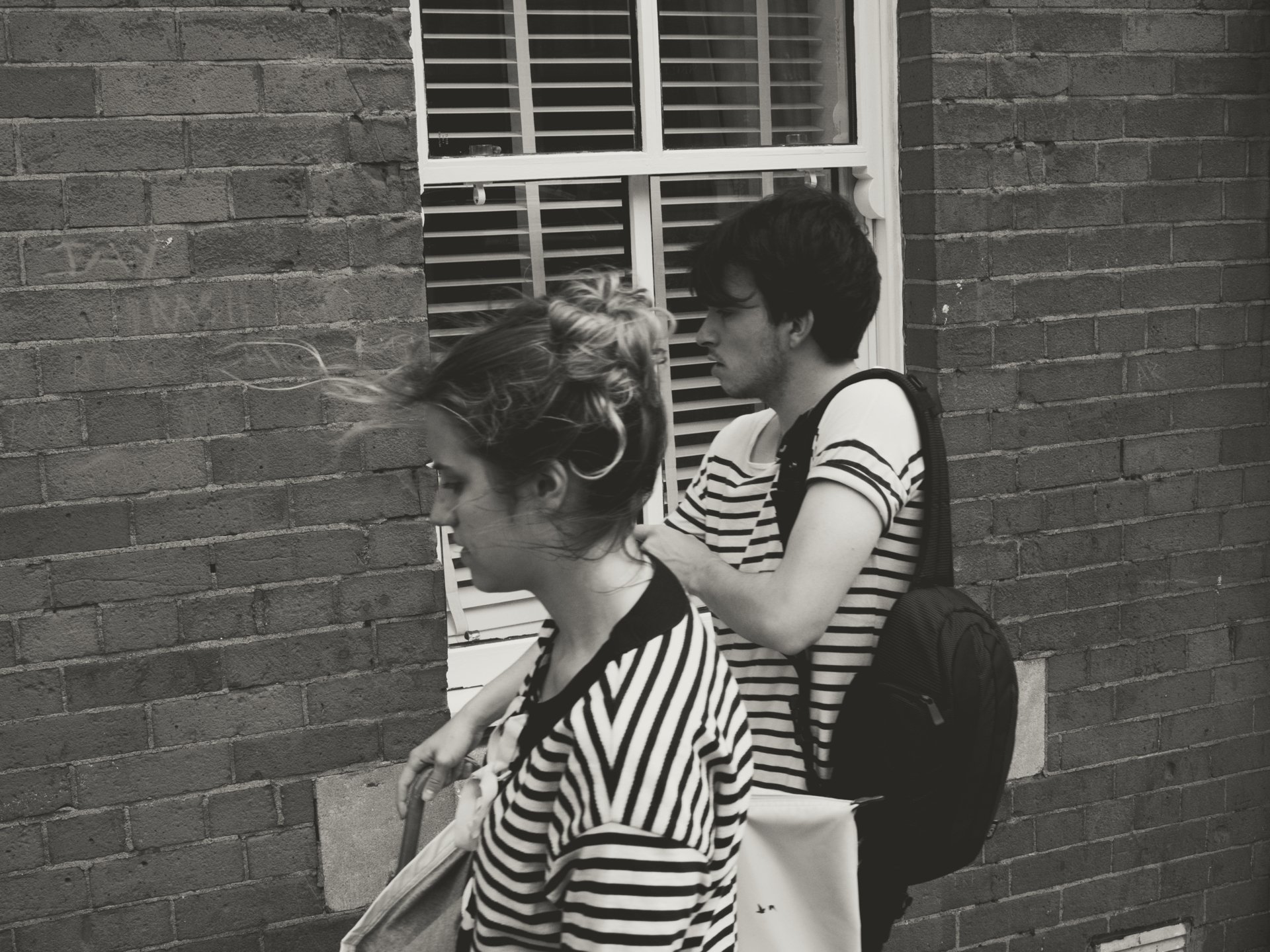A young woman and a young man are walking on a sidewalk against a brick wall. The woman has windblown hair and is wearing a striped shirt. The man has dark hair, a striped shirt, and is carrying a backpack. The woman is pushing a shopping cart.