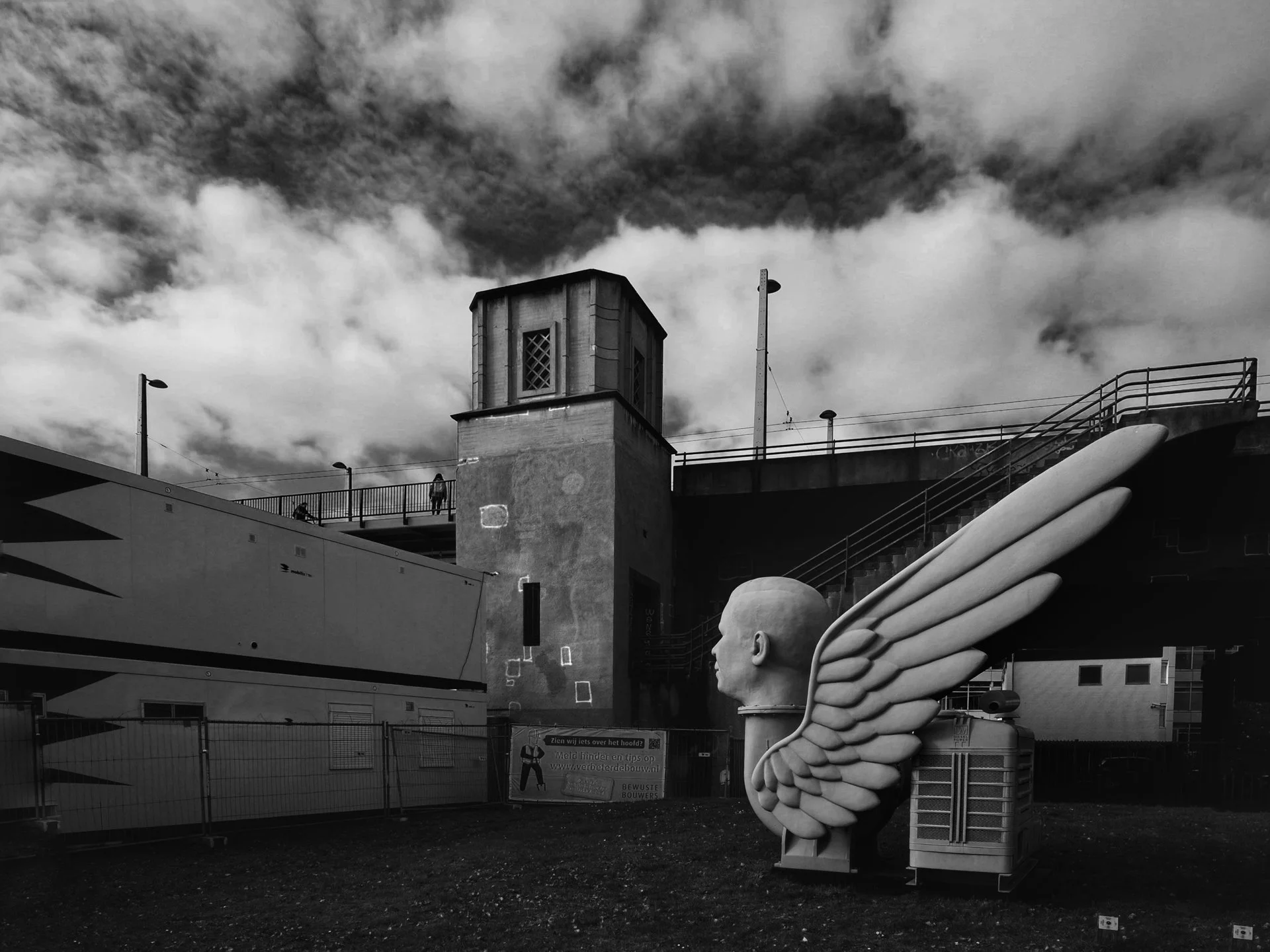 Black and white photo of a public space with a sculpture of a human head with large wings attached to the back. The sculpture is situated outdoors near a fence. In the background, there is a bridge or elevated walkway with a few people walking and so