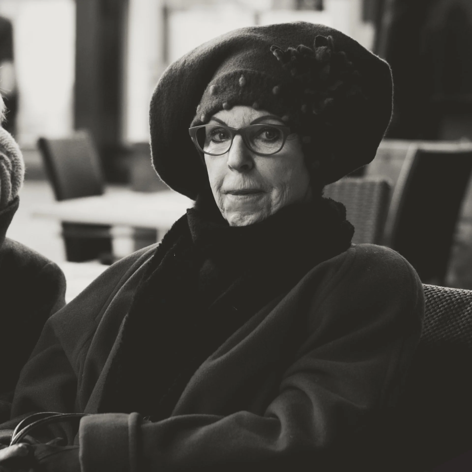 Close-up black and white photo of a woman with glasses, wearing a large hat, sitting indoors.
