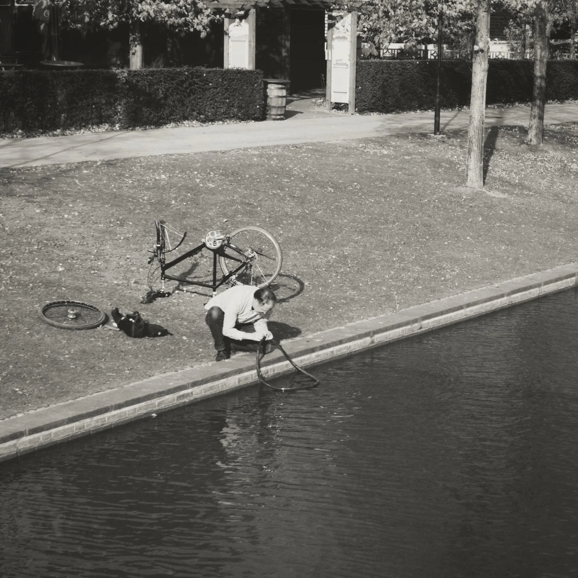 A flooded street with a bicycle on the sidewalk, a person crouching near the curb, and a house in the background with a hedge and trees.