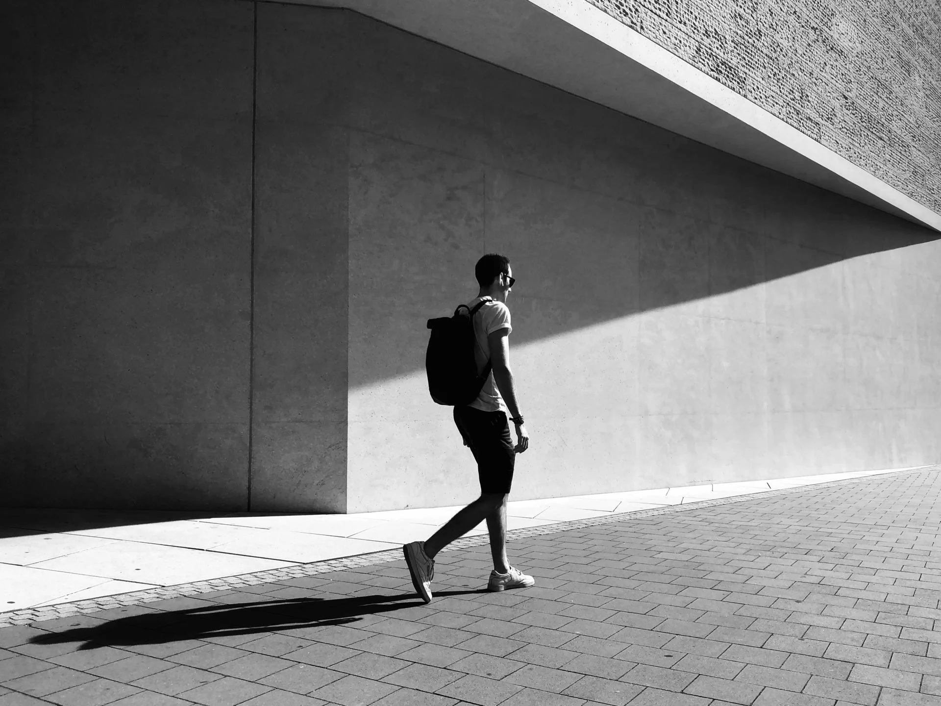 A young man with a backpack and sunglasses walking on a brick pavement against a plain wall. Shadows cast on the ground and wall in bright sunlight.